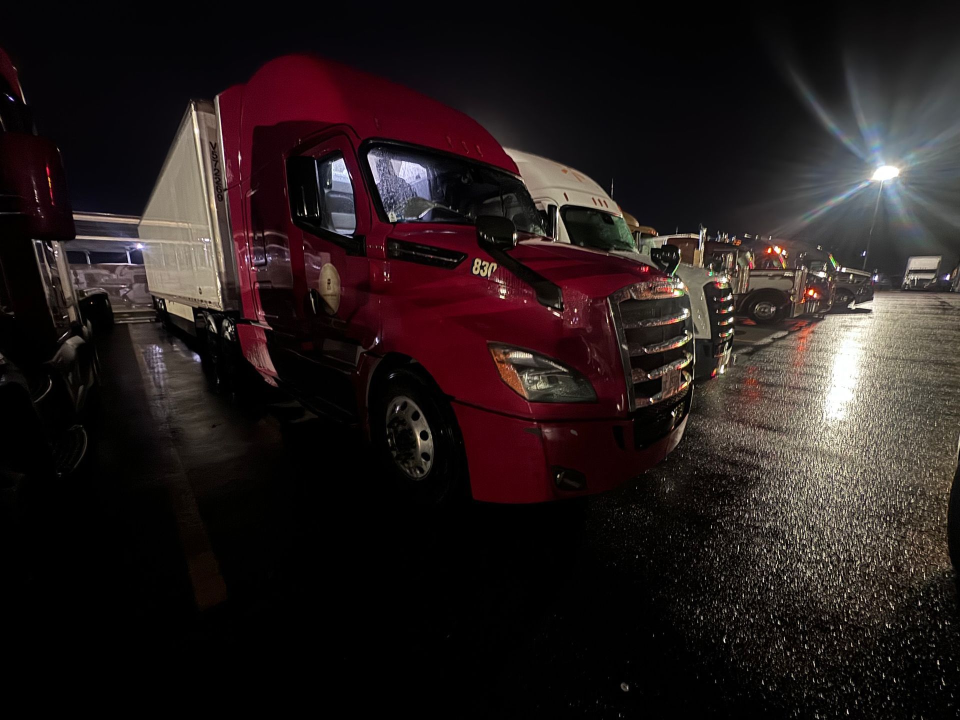 Red semi-truck parked in a wet lot at night, other trucks in the background.