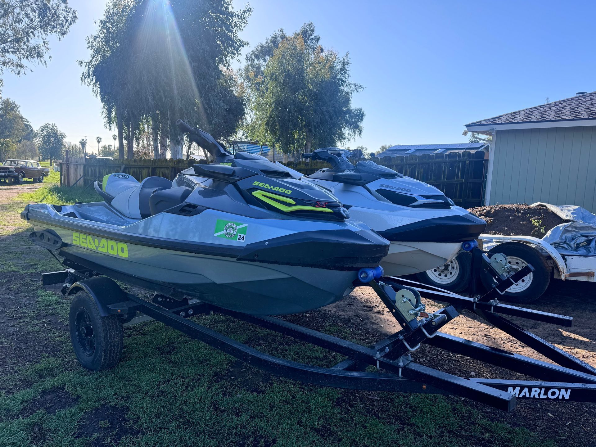 Two Sea-Doo jet skis on a trailer, parked on grass in front of a house.