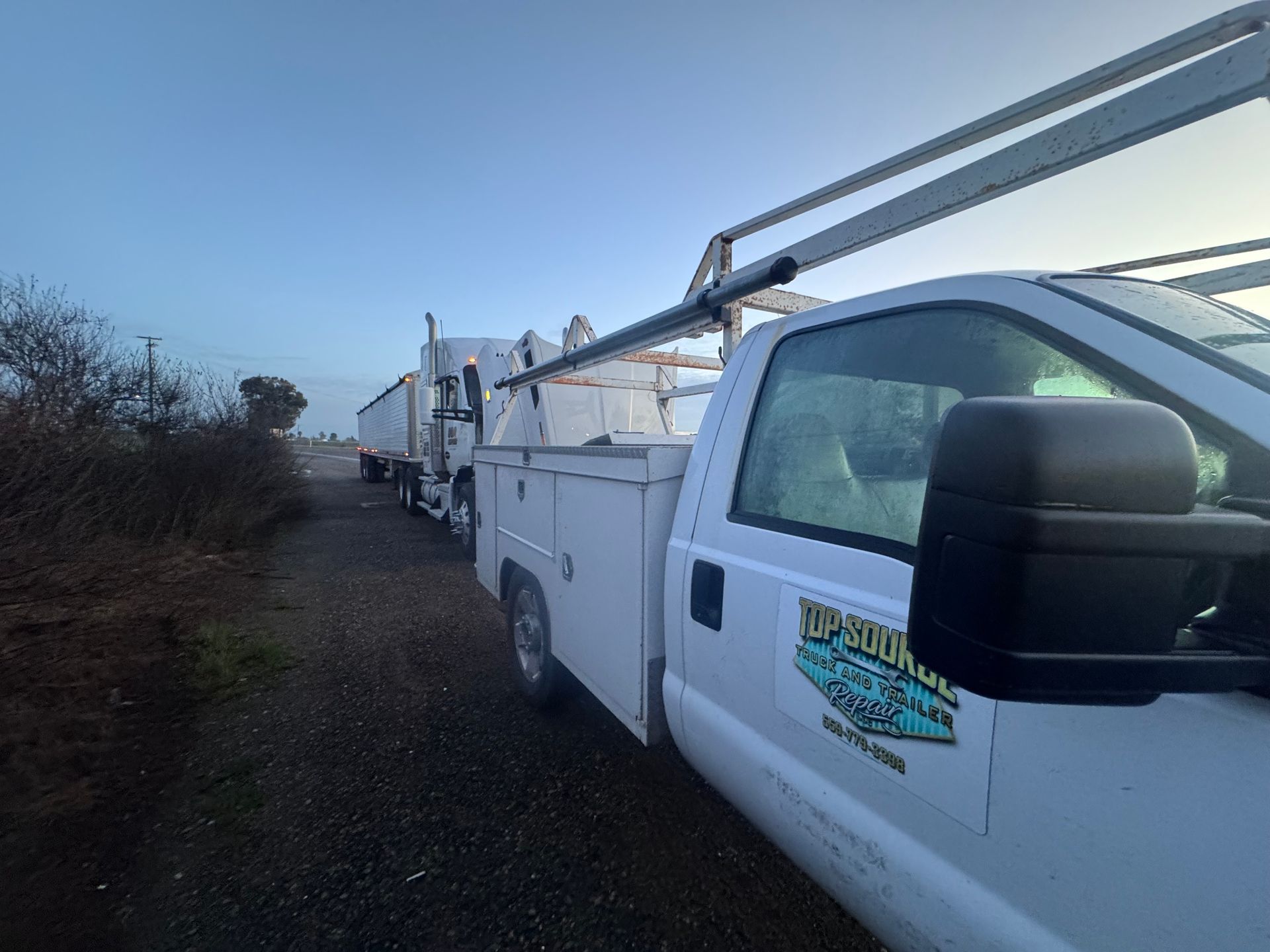 White work truck with ladder rack, parked next to a truck with a trailer.