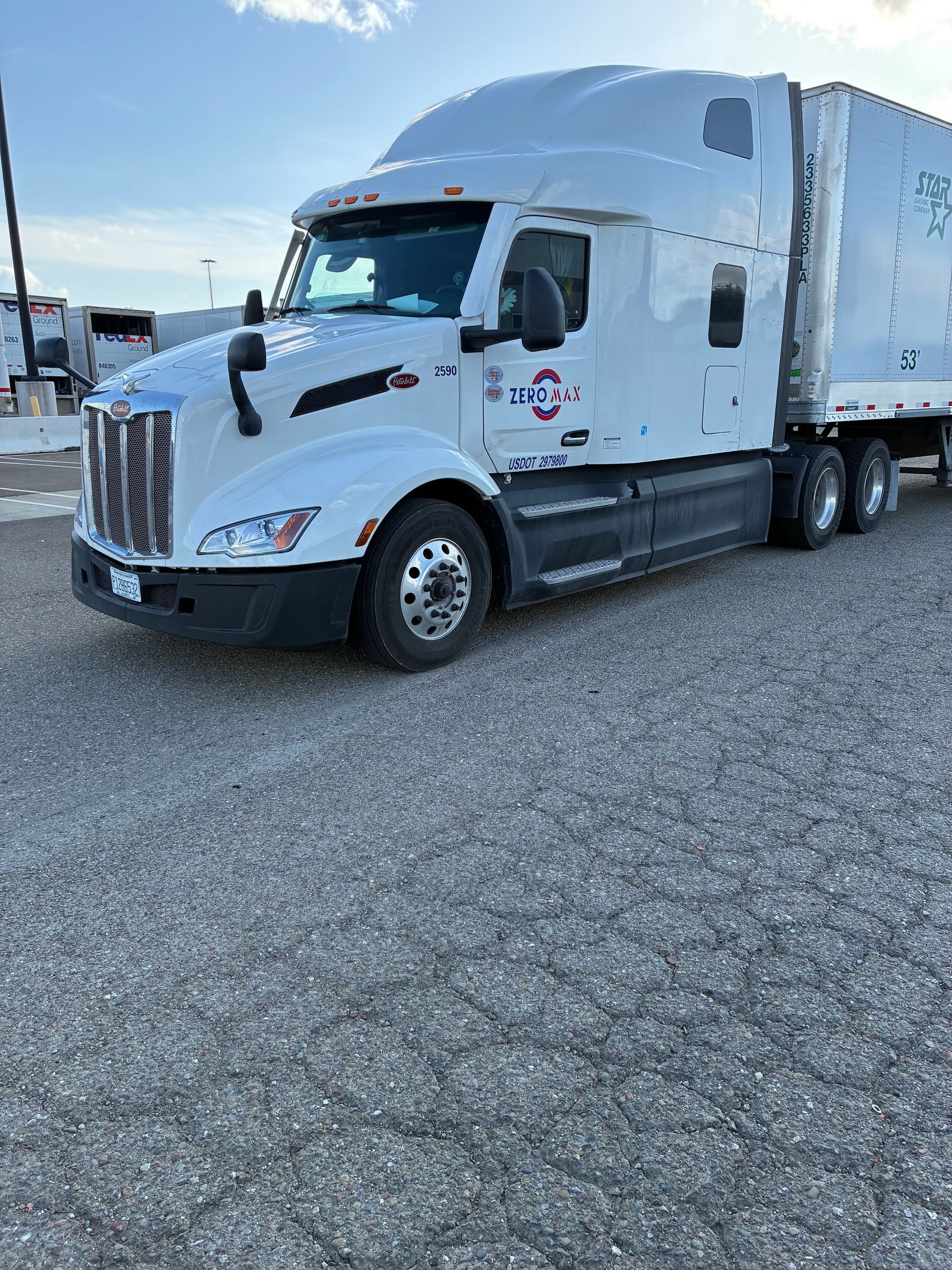White semi-truck parked on gravel. Daytime, blue sky. Side view of cab and trailer with company logo.