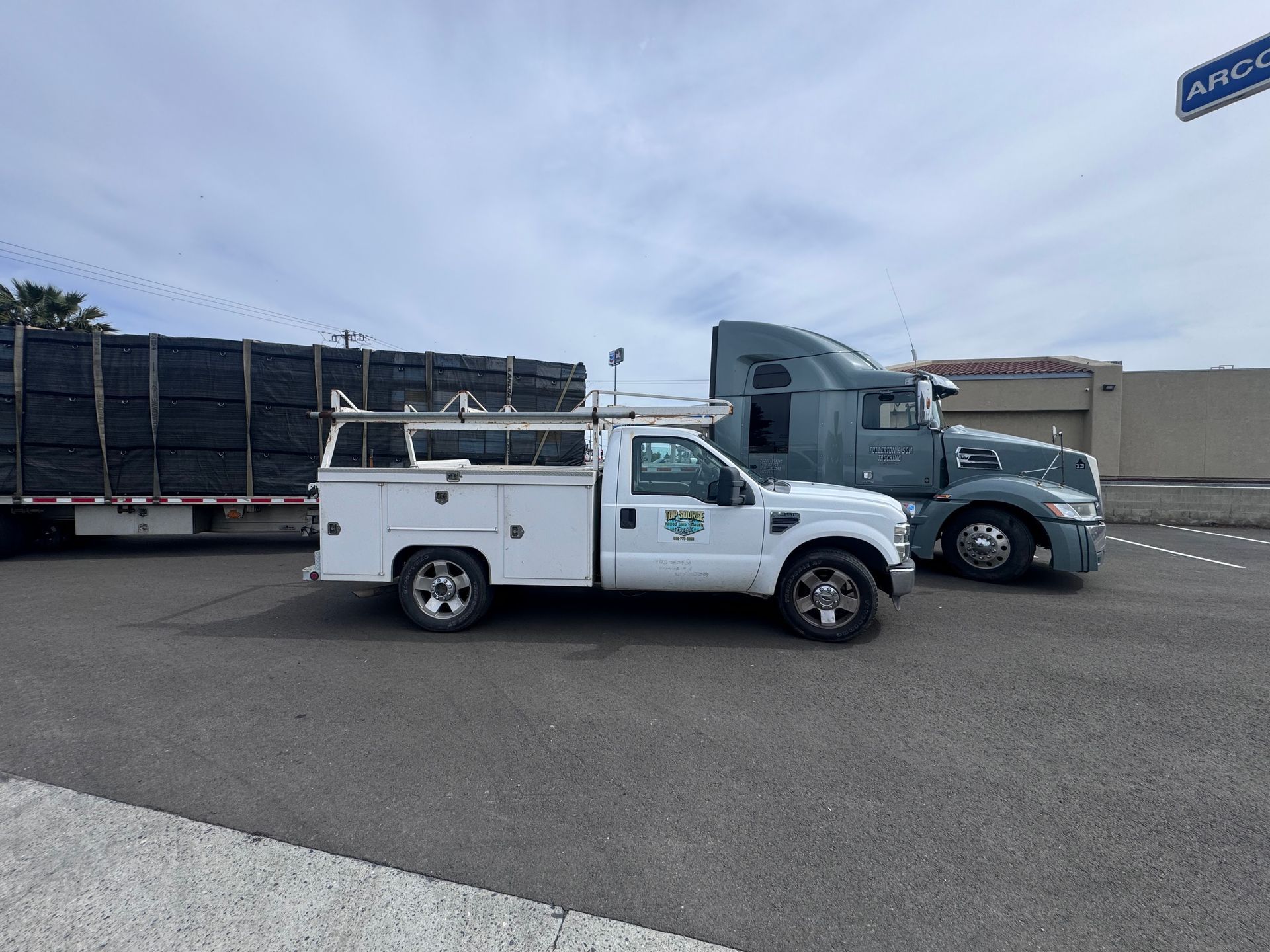 White work truck parked next to a semi-truck, with a flatbed trailer loaded with black materials. Outdoors.