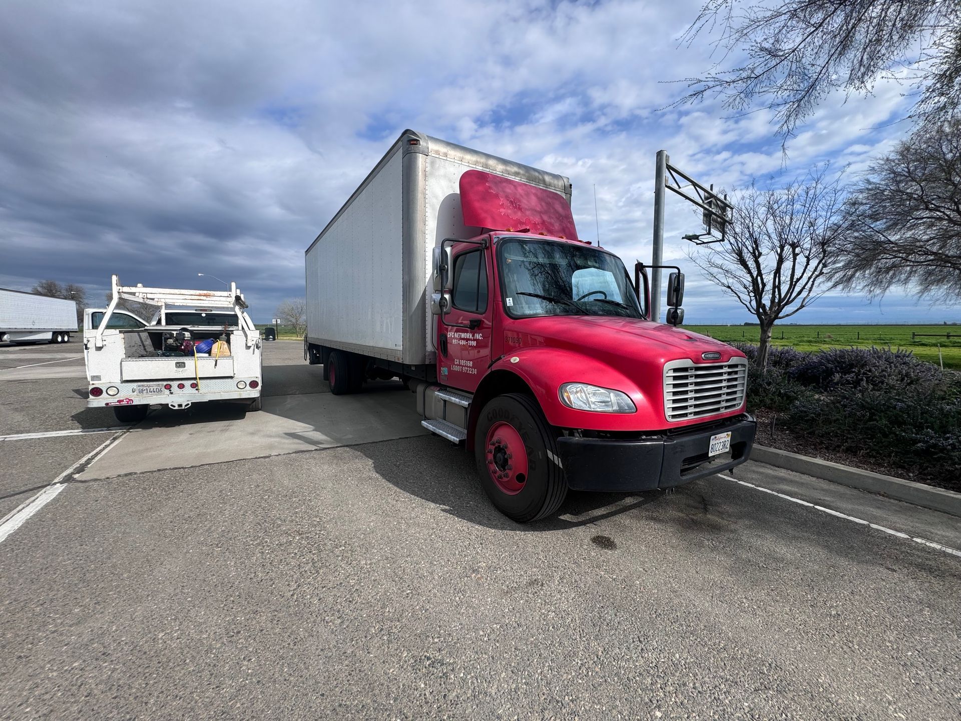 Red box truck parked on asphalt; white utility truck to the left, under a cloudy sky.