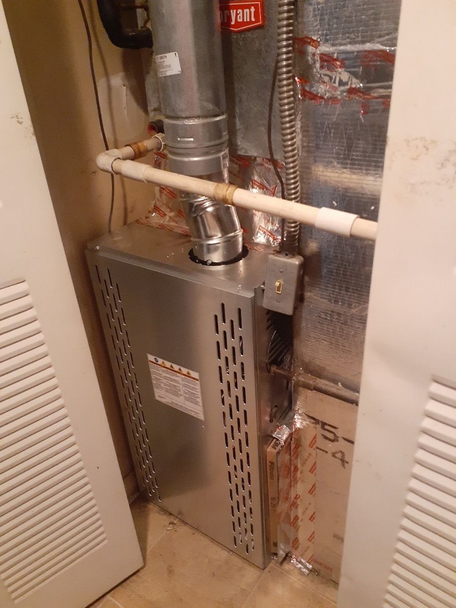 Furnace enclosed in a narrow space, with metal vents and ductwork. White pipes run horizontally.