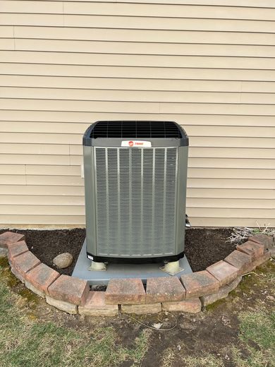 Gray Trane air conditioner unit set on a concrete pad and surrounded by brick near a beige wall and grass.