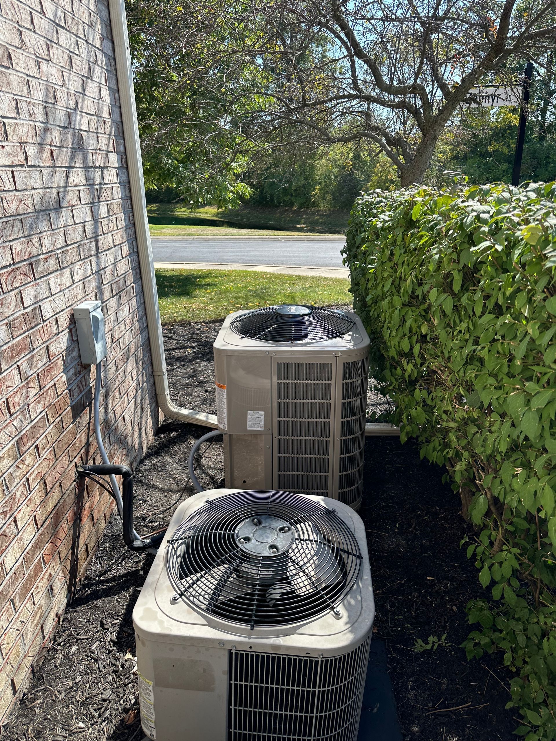 Two beige air conditioning units sit near a brick wall and green hedge outside.