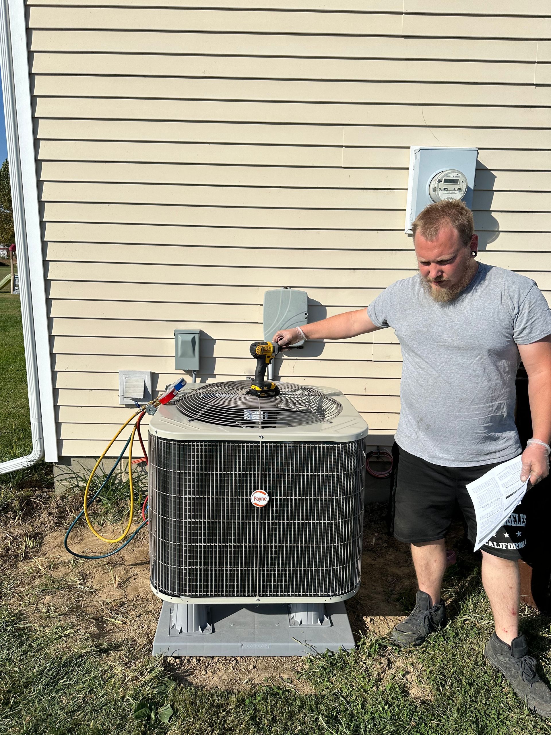 Man in shorts using a drill on an AC unit outdoors near a house.