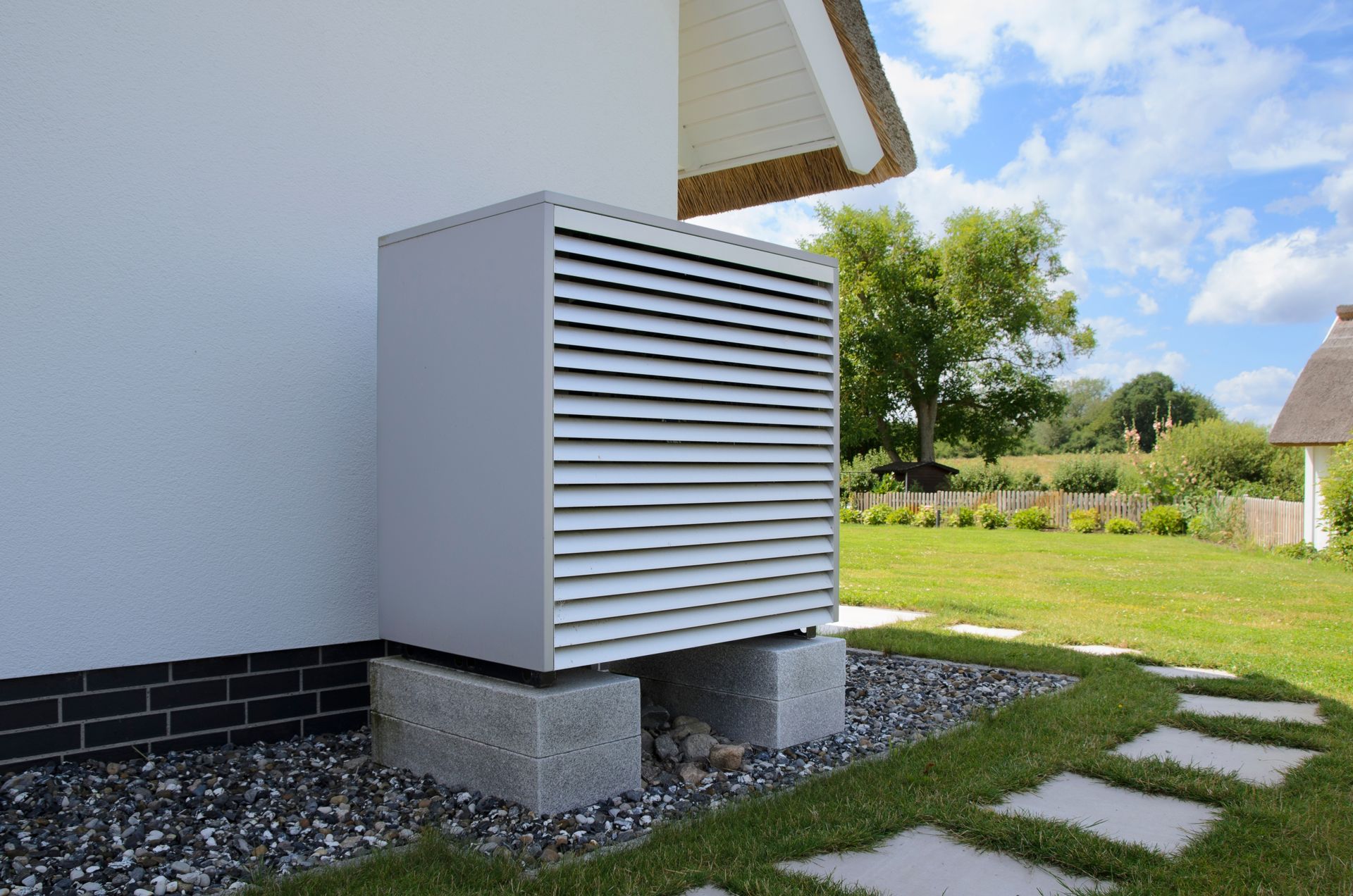 Exterior view of a heat pump on concrete blocks in a grassy yard, beside a white house.