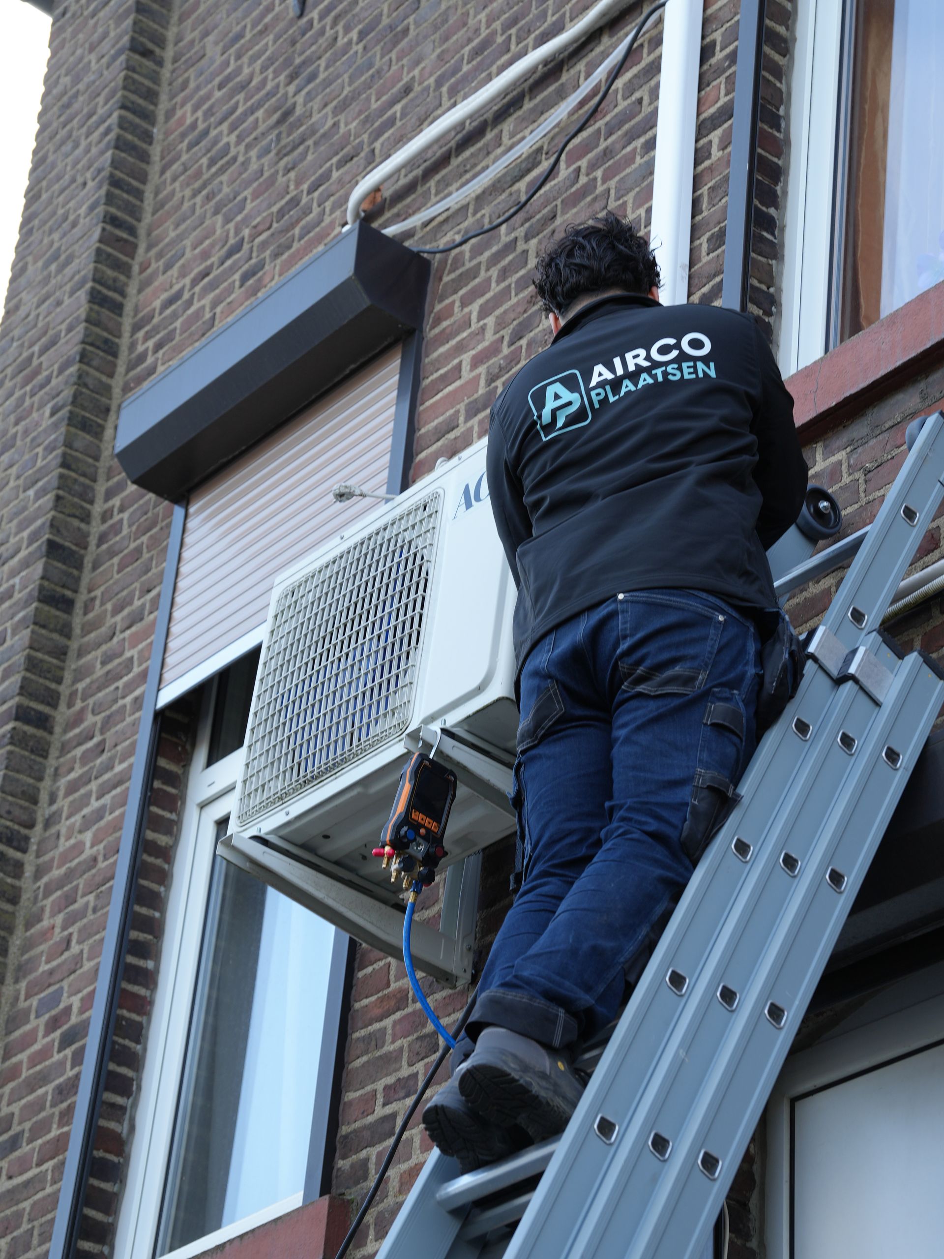 Technicus op ladder bezig met het repareren van een aan de muur gemonteerde airconditioner in een bakstenen gebouw.