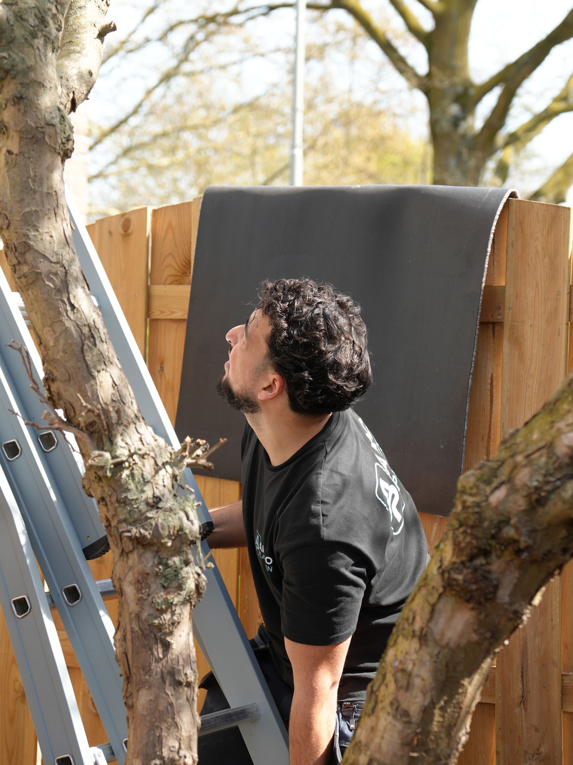 Een man staat buiten naast een boom en kijkt omhoog naar een groot grijs paneel dat tegen een houten schutting leunt.