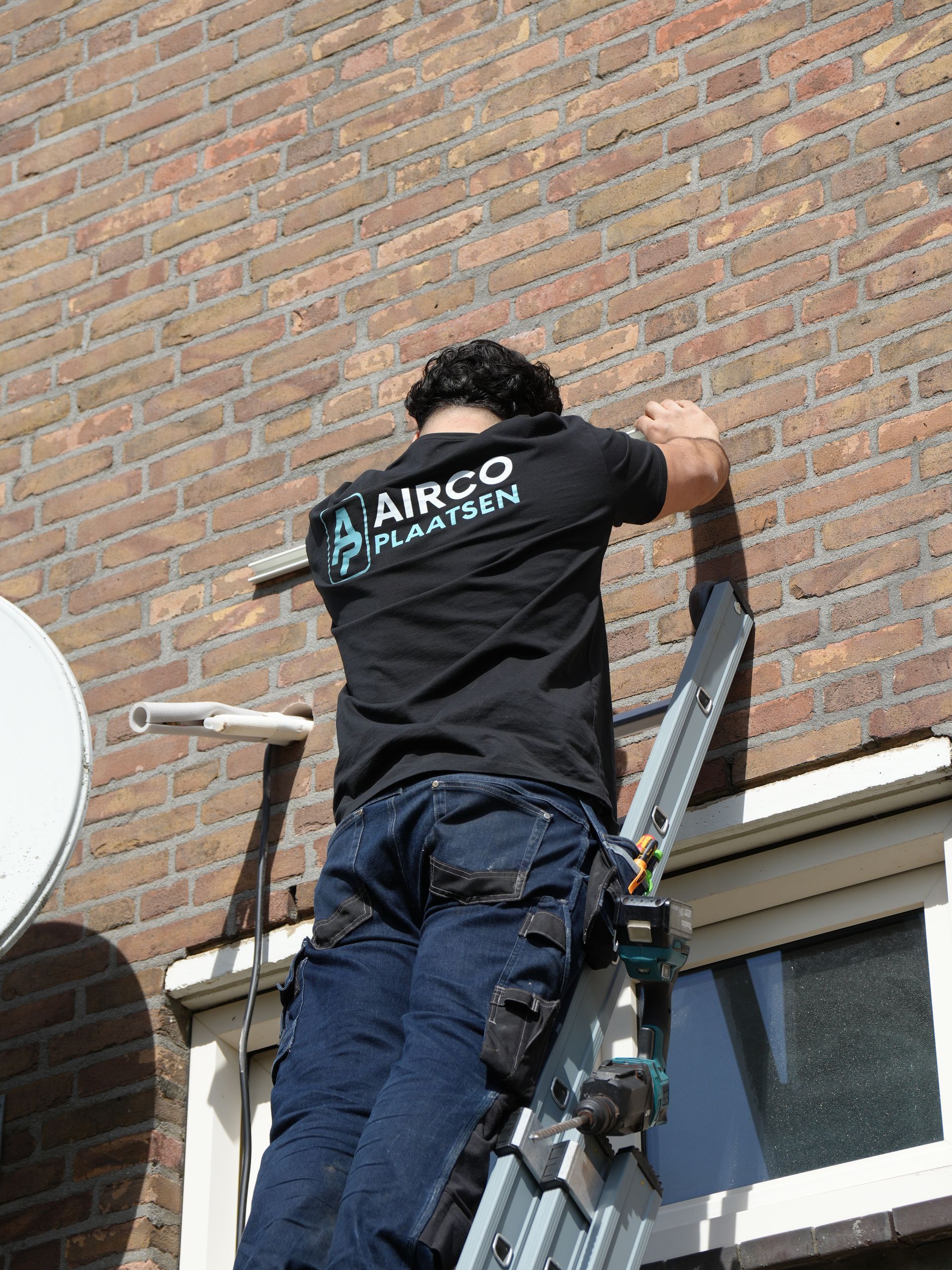 Een arbeider in een zwart shirt gebruikt een ladder om een ​​bakstenen muur boven een raam te repareren.