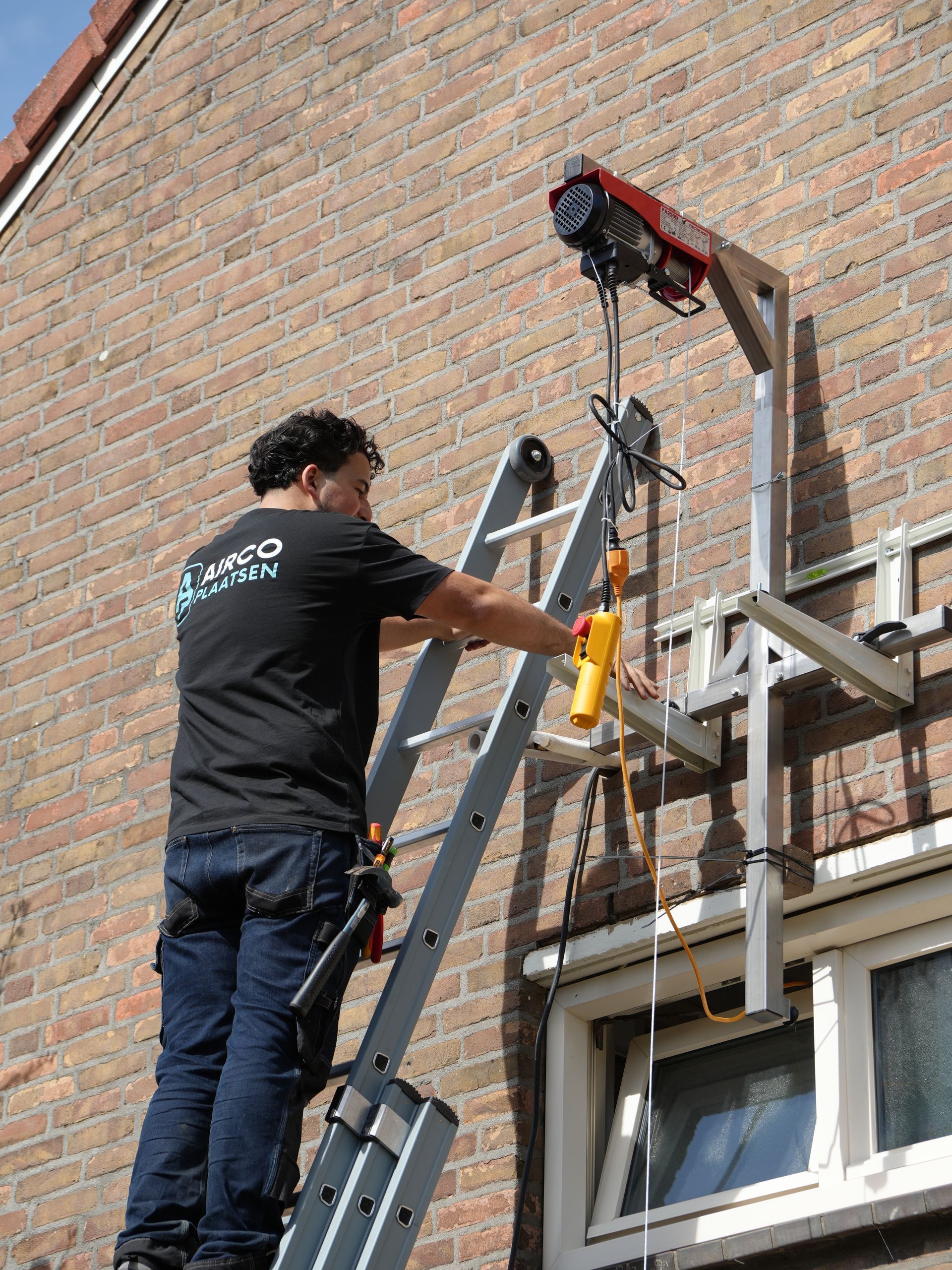 Een werknemer gebruikt een ladder en een takel om apparatuur tegen een bakstenen muur boven een raam te tillen.