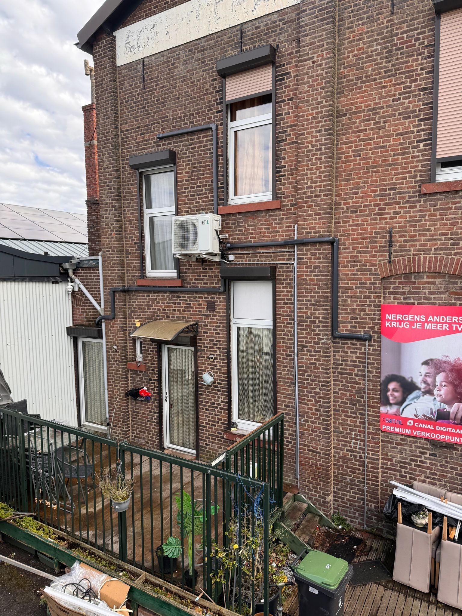 Gevel van een appartementencomplex van baksteen met ramen, airconditioning en een kleine omheinde tuin eronder.