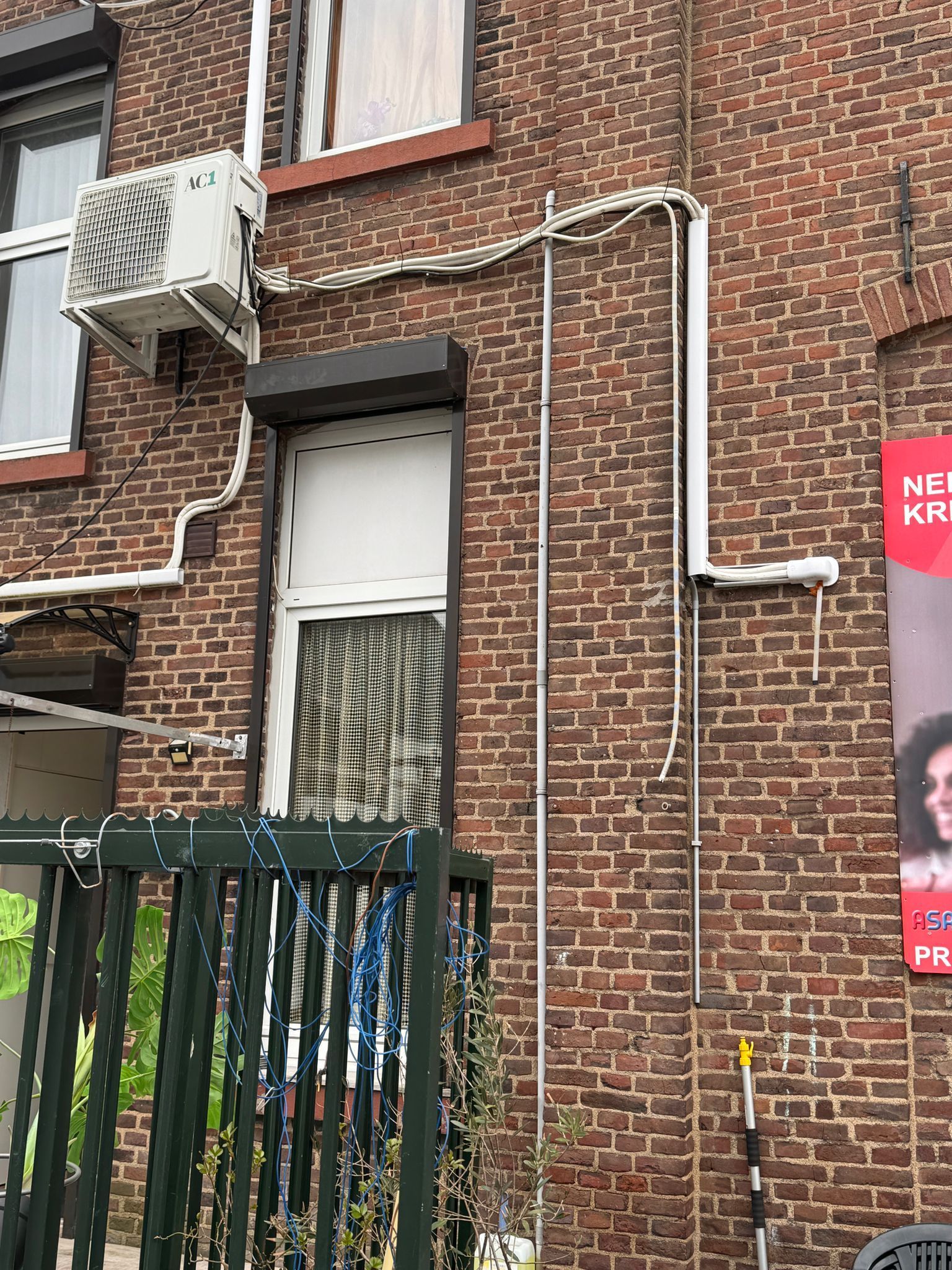 Bakstenen gevel met wit omlijste ramen, een aan de muur gemonteerde airconditioner en een roze poster aan de rechterkant.