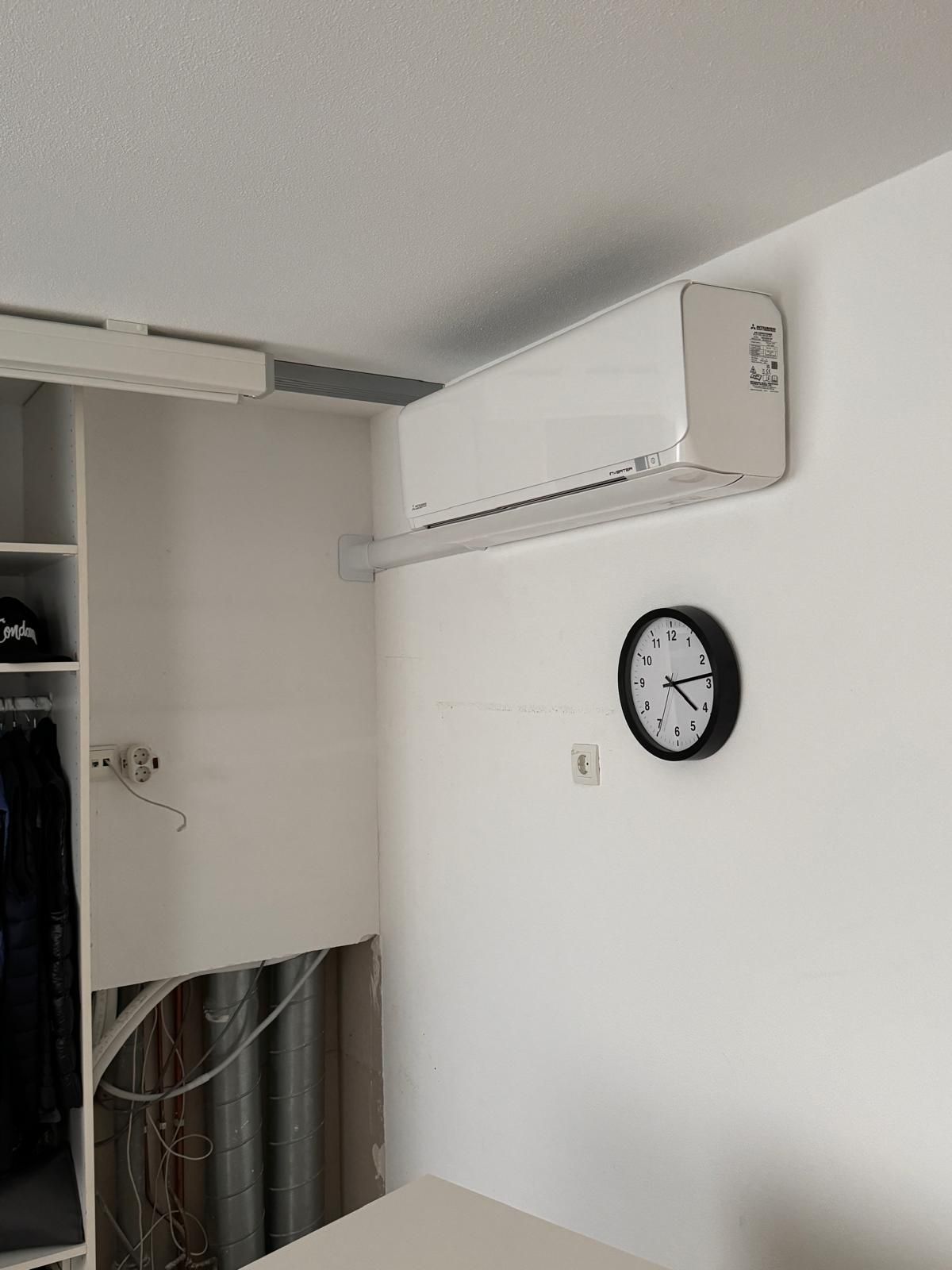 Witte kamerwand met gemonteerde airconditioner en een zwarte klok boven een open kast.