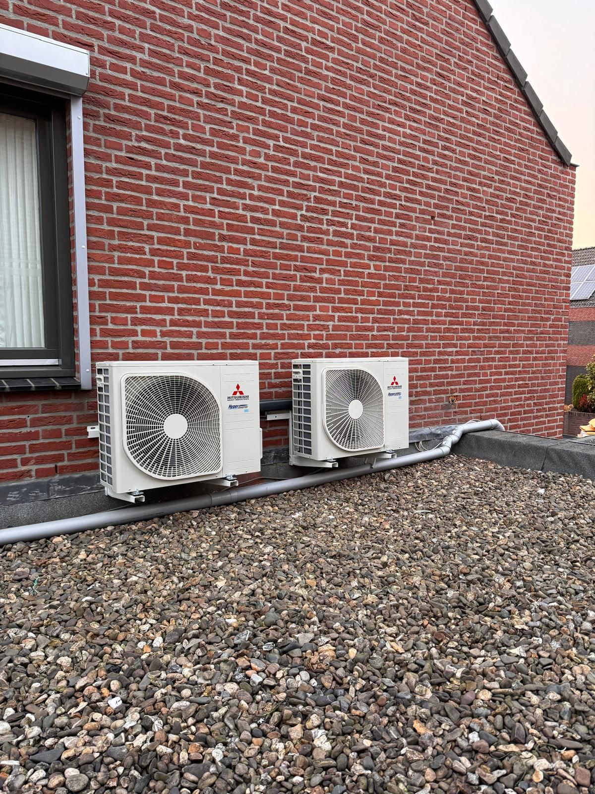 Twee witte buitenunits voor de airconditioning staan naast een rood bakstenen huis met een grinddak aan de voorkant.