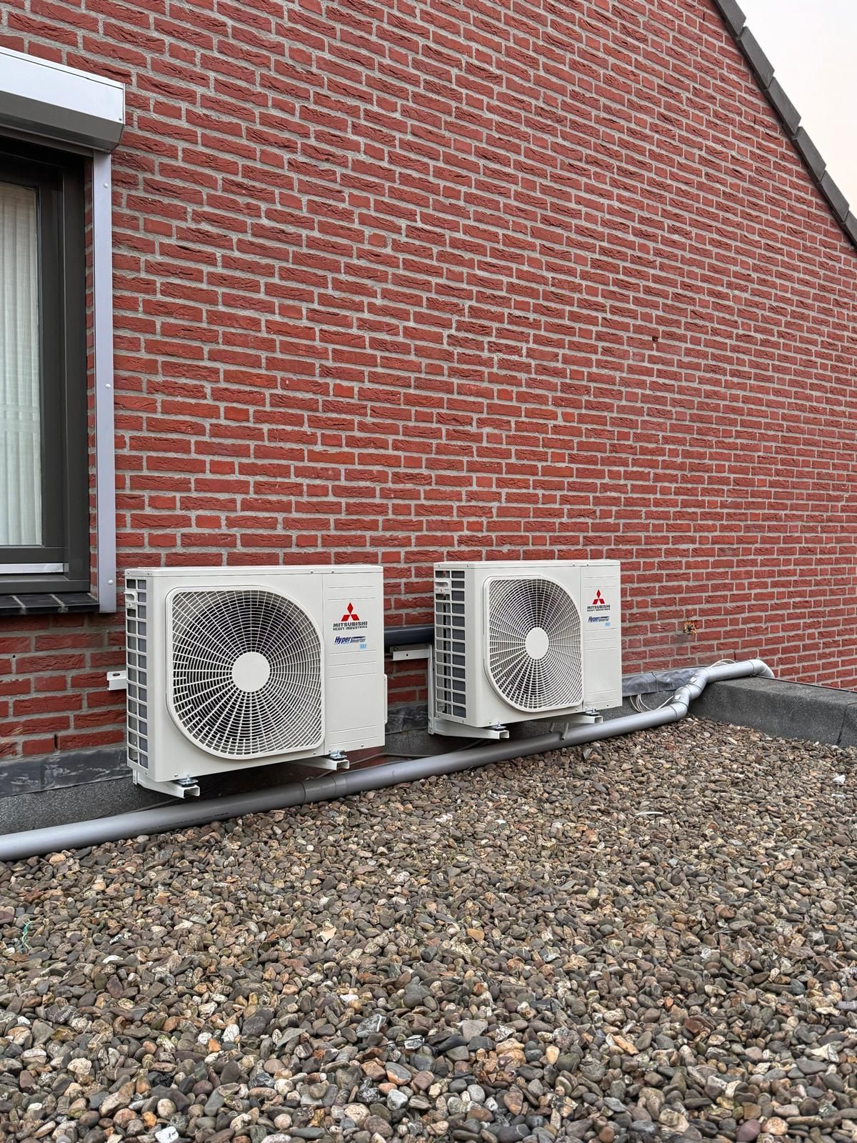 Twee witte buitenunits voor de airconditioning staan naast een rode bakstenen muur op een met grind bedekte ondergrond.