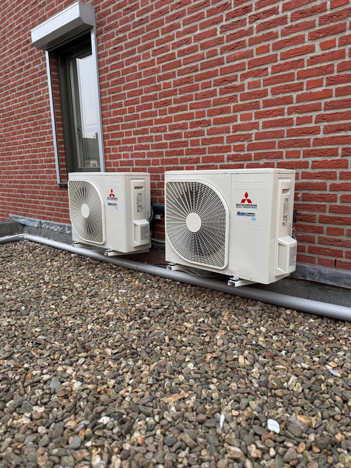 Twee Mitsubishi Electric-buitenunits staan naast een bakstenen gebouw op een grindbed.