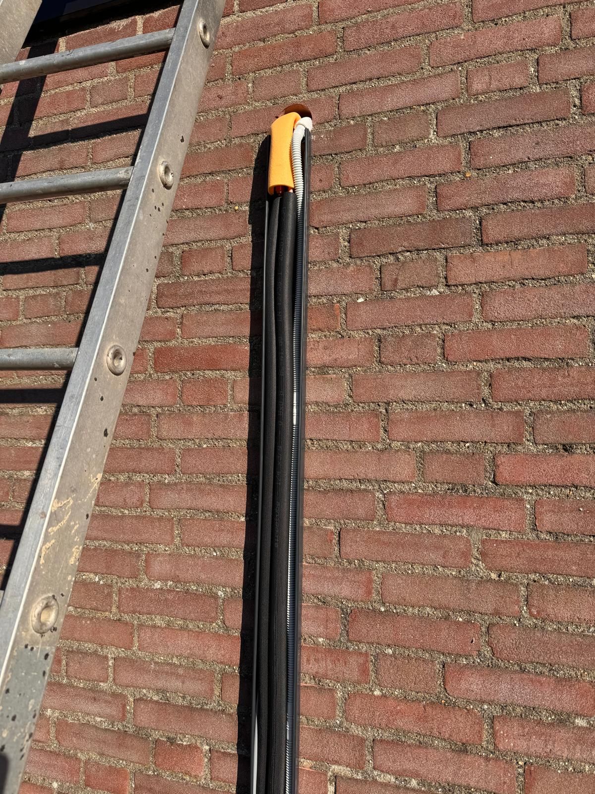Zwarte kabels hangen aan een gele beugel aan een bakstenen muur naast een metalen ladder.