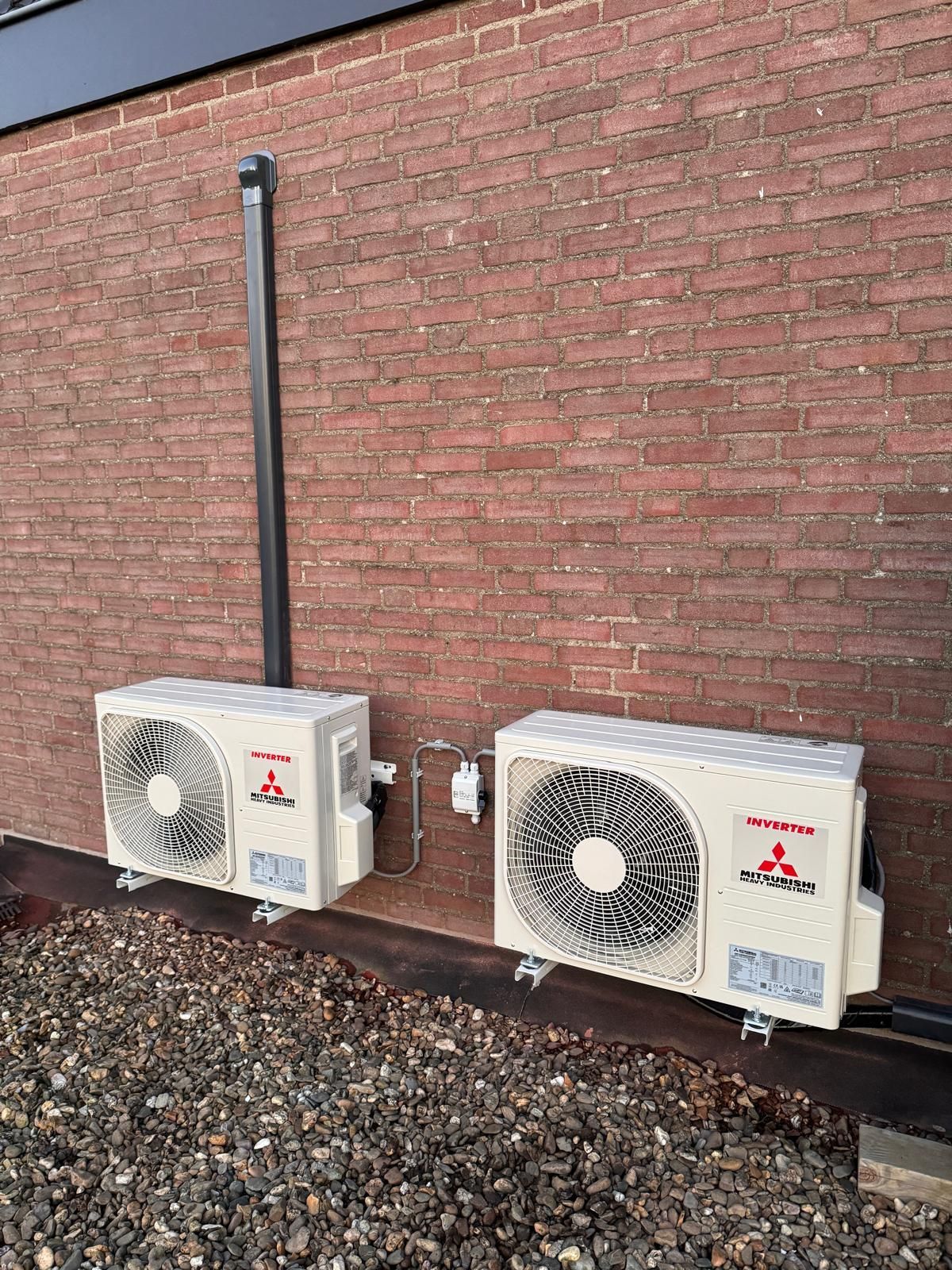 Twee witte HVAC-units gemonteerd naast een bakstenen muur met grind eronder.