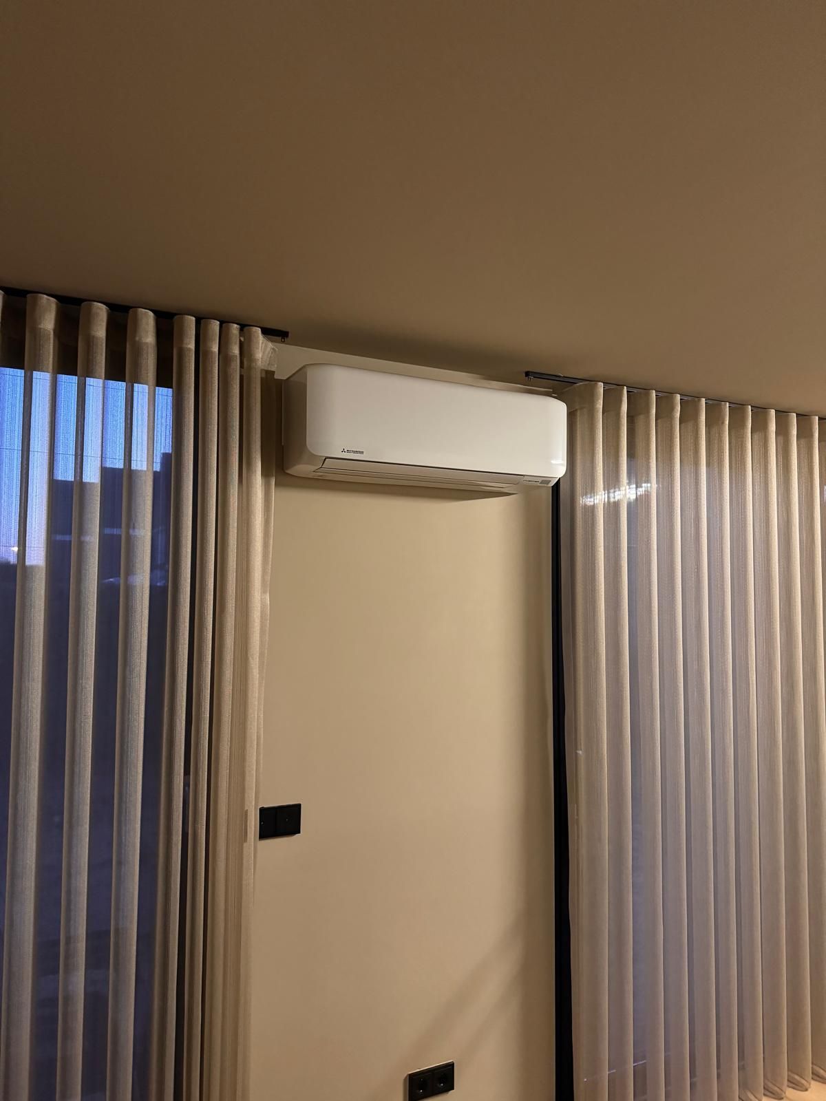 Wandgemonteerde airconditioner boven een beige muur tussen twee hoge verticale jaloezieën in de schemering.