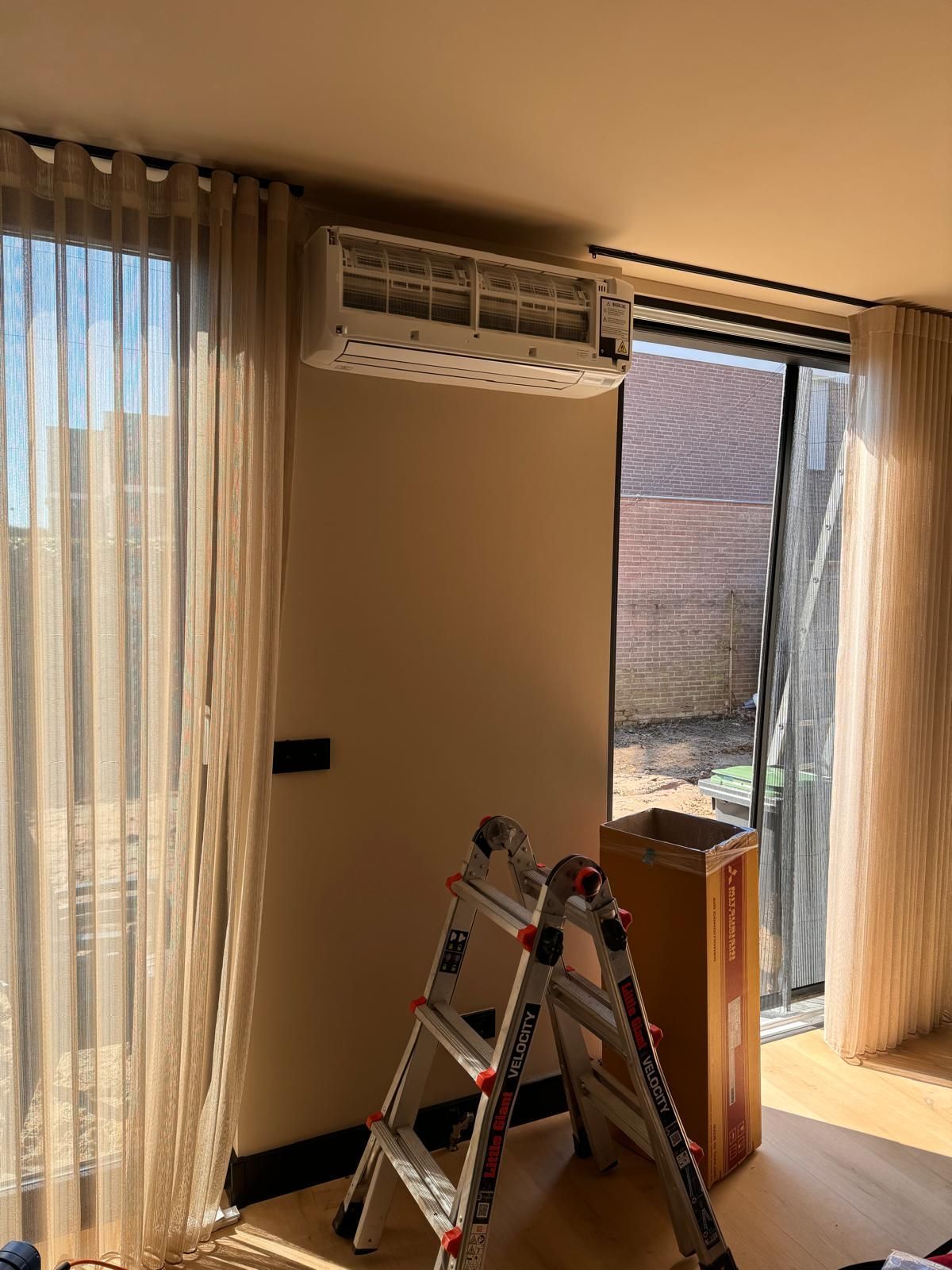 Woonkamer met ladder, openslaande schuifdeur, vitrage en aan de muur gemonteerde airconditioning.