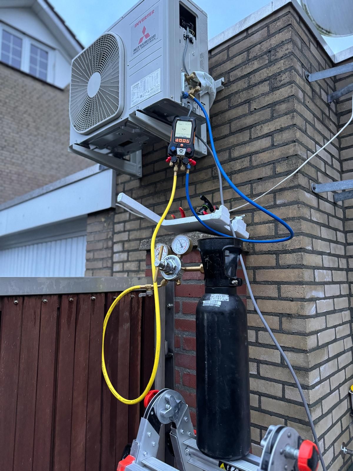 Buitenunit van de airconditioning, gemonteerd boven een zwarte koelmiddeltank met kleurrijke slangen aan een bakstenen muur.