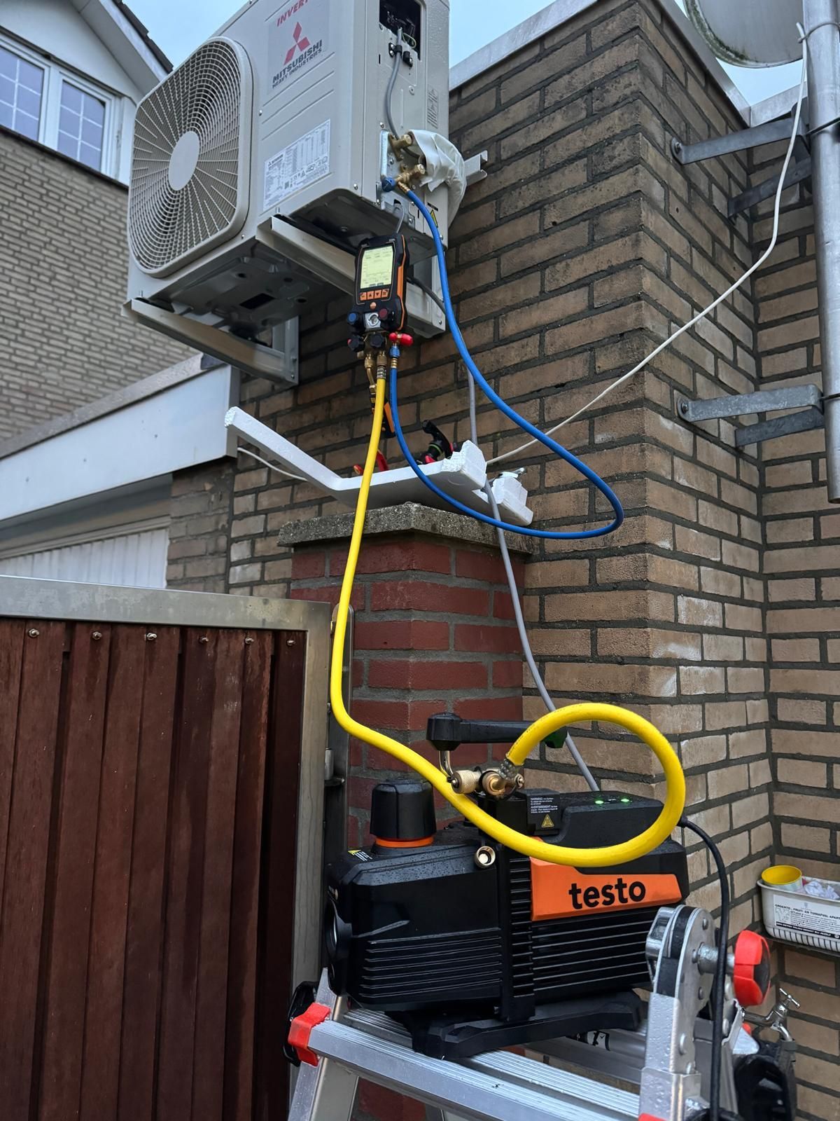 Gele slang aangesloten op de HVAC-unit aan de buitenmuur van baksteen, met daaronder testapparatuur.