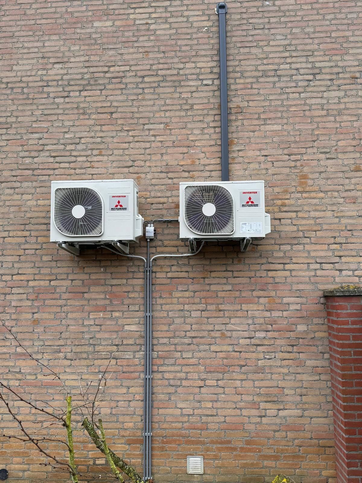 Twee aan de muur gemonteerde airconditioningunits op een bakstenen buitenmuur met zichtbare leidingen.
