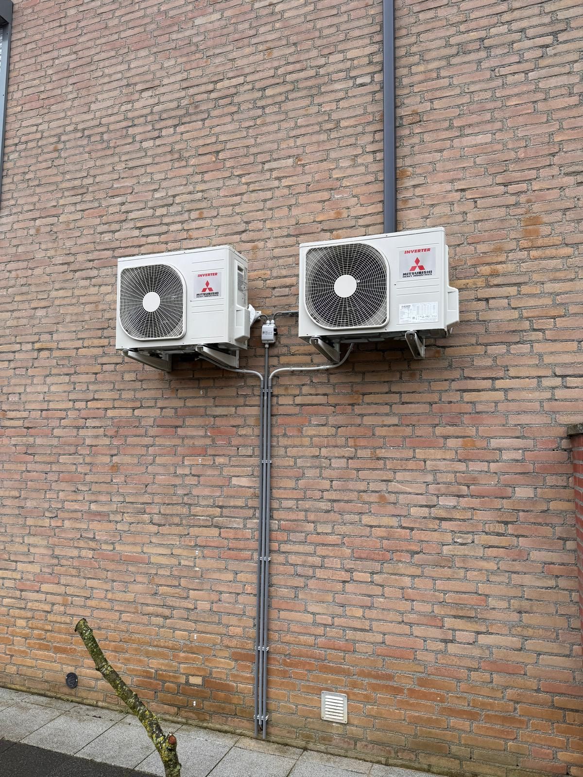 Twee buitenunits voor airconditioning, gemonteerd op een bakstenen muur met zichtbare leidingen.