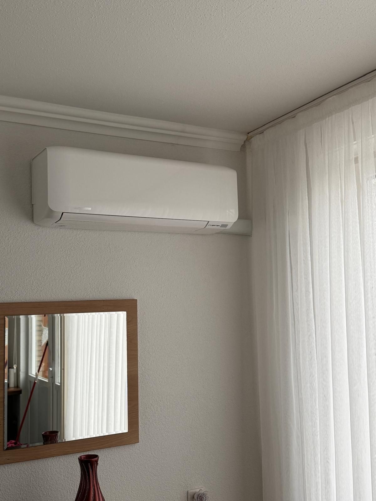 Witte, aan de muur gemonteerde airconditioner boven een spiegel en een doorschijnend gordijn in een lichte kamer.