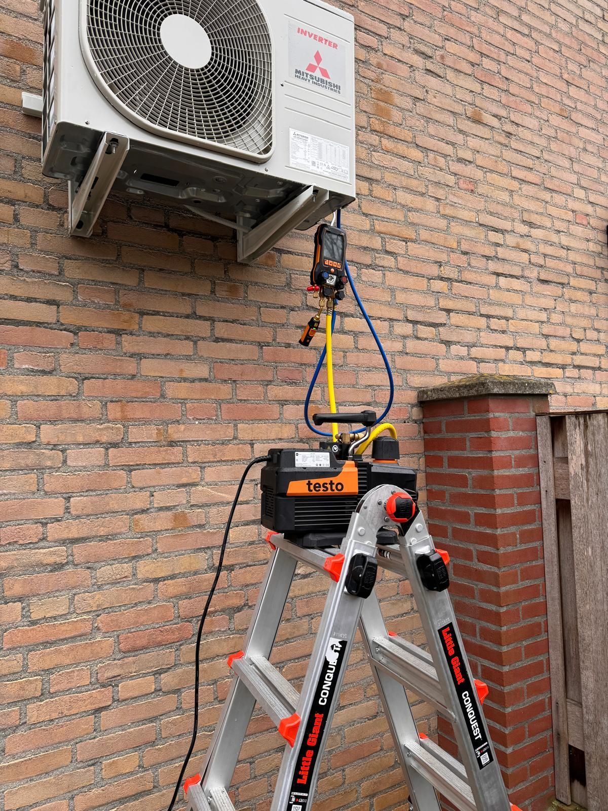 Een airconditioningunit wordt gerepareerd aan een bakstenen muur, met een ladder en gereedschapstas eronder.