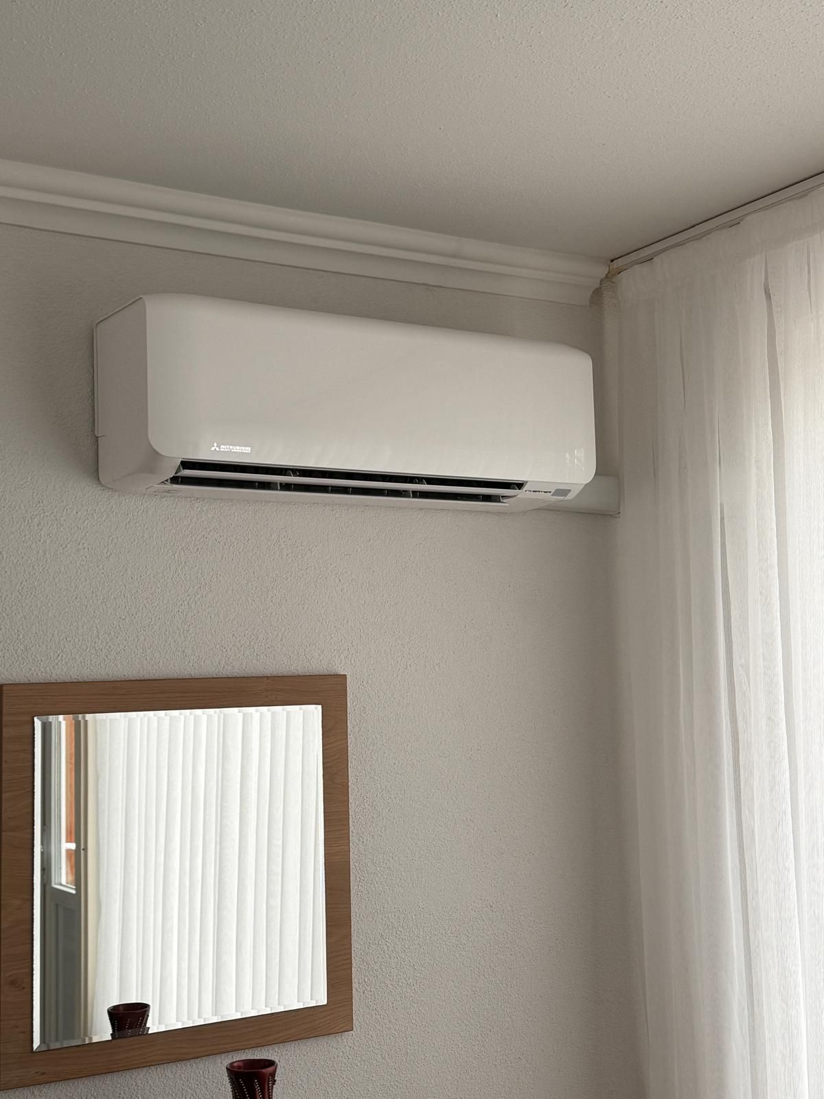 Een aan de muur gemonteerde witte airconditioner boven een spiegel met houten frame in een beige kamer.