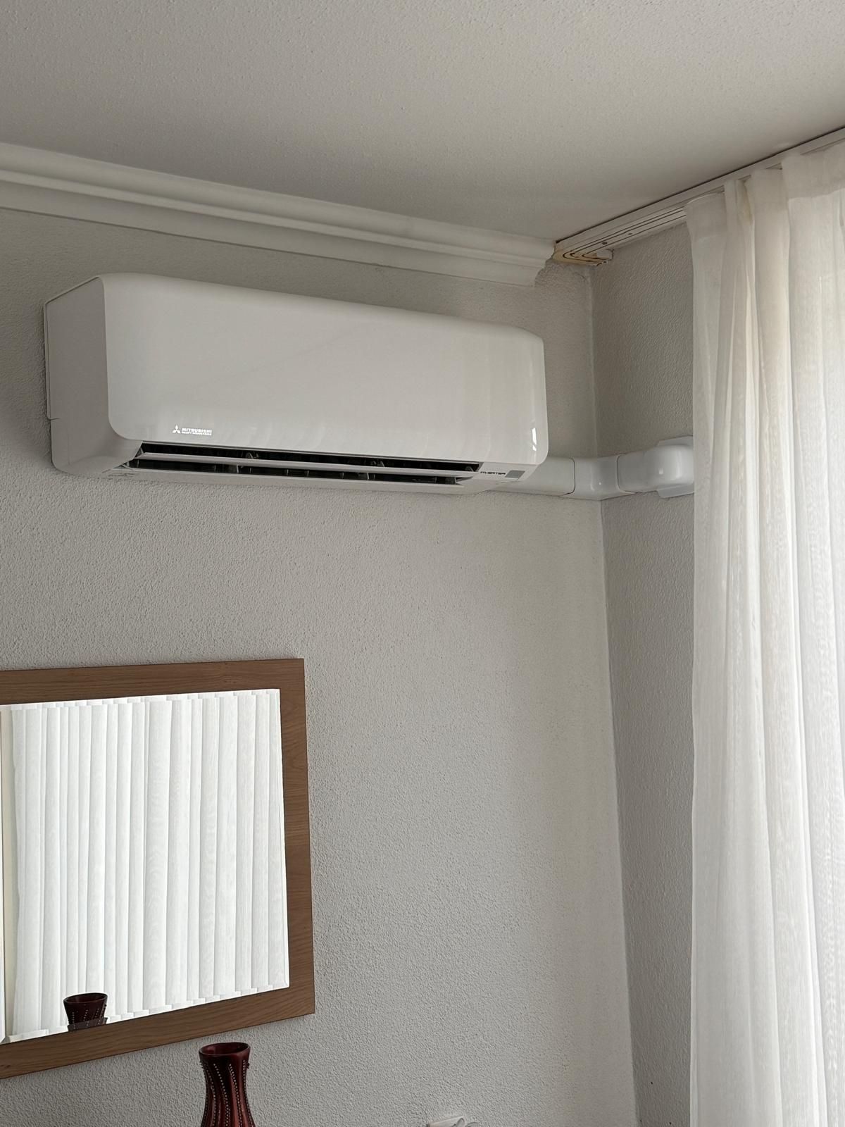 Een aan de muur gemonteerde witte airconditioner boven een spiegel in een lichtgrijze kamer met een doorschijnend gordijn.