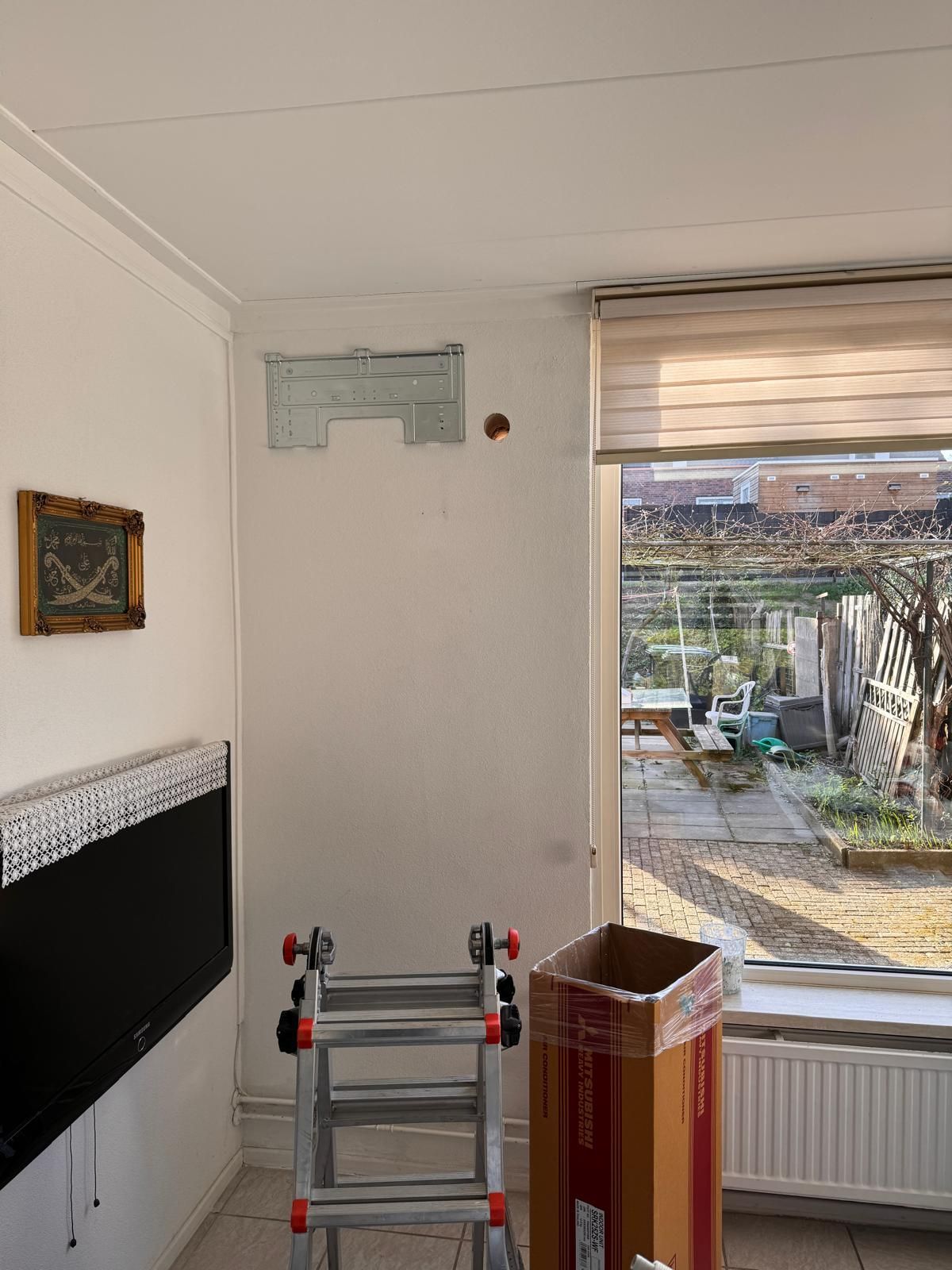 Woonkamerhoek met ladder, tv aan de muur, open raam, radiator en gestapelde dozen bij het raam.