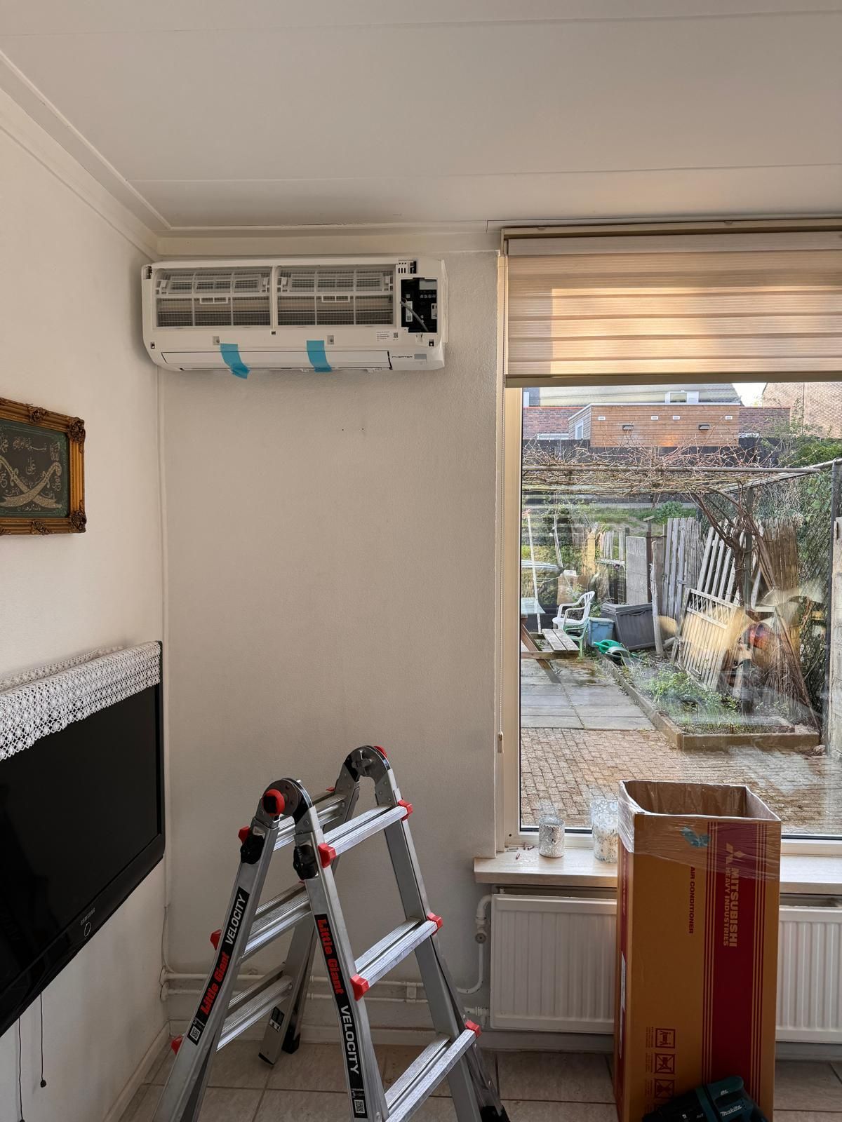 Woonkamer met aan de muur gemonteerde airconditioning, ladder, tv en kartonnen doos bij een raam met uitzicht op een patio.