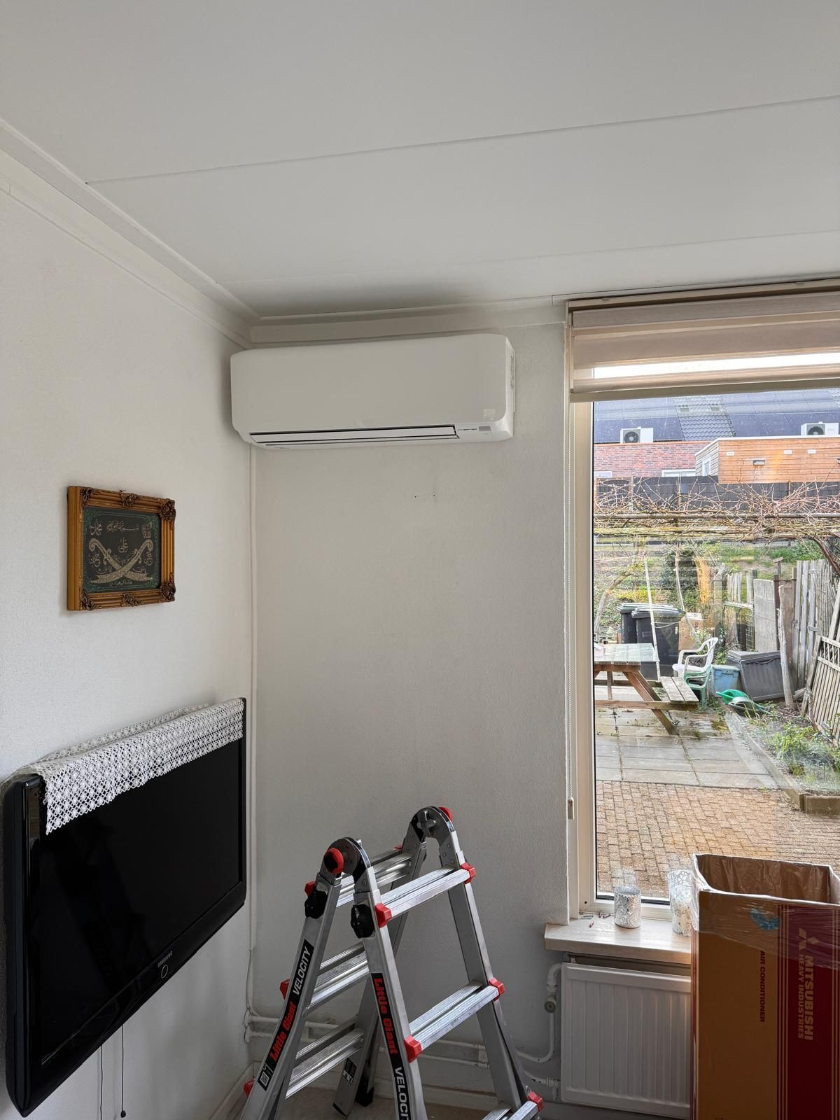 Kleine kamer met aan de muur gemonteerde tv, airconditioning, ladder en een open raam met uitzicht op de tuin.