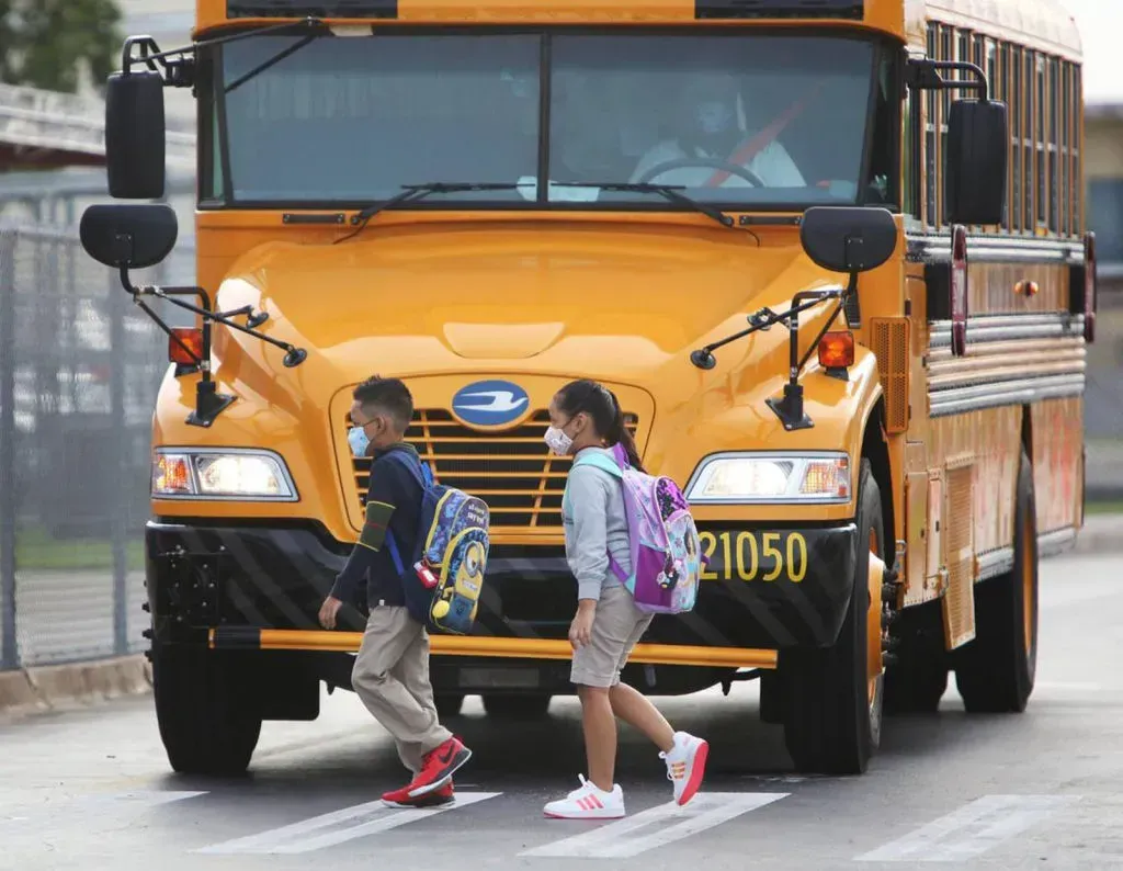Two children wearing masks are crossing the street in front of a school bus.