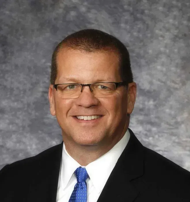 A man in a suit and tie is smiling in front of a wooden wall.