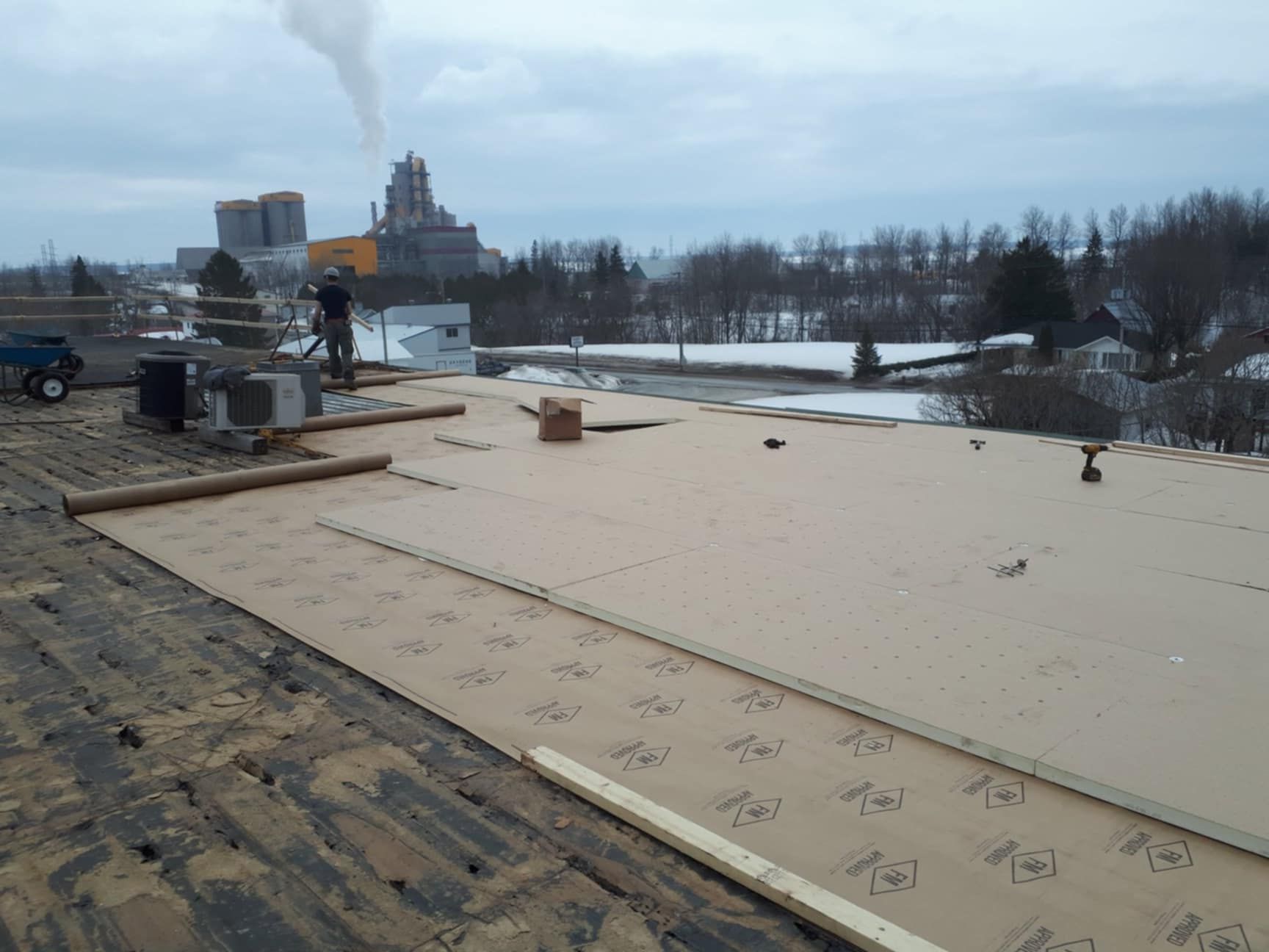 Des ouvriers du bâtiment installent de l'isolant sur un toit plat. Neige et bâtiments industriels en arrière-plan. Journée nuageuse.