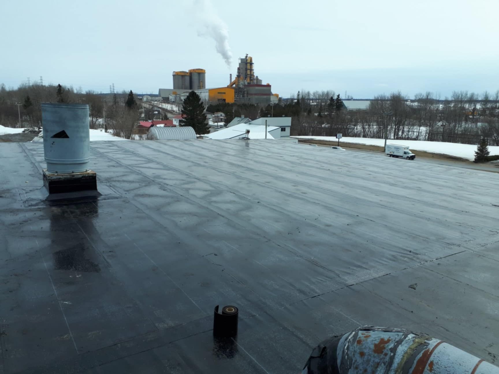 Vue aérienne d'un toit plat noir avec des tuyaux, d'un complexe industriel lointain et d'un sol recouvert de neige.