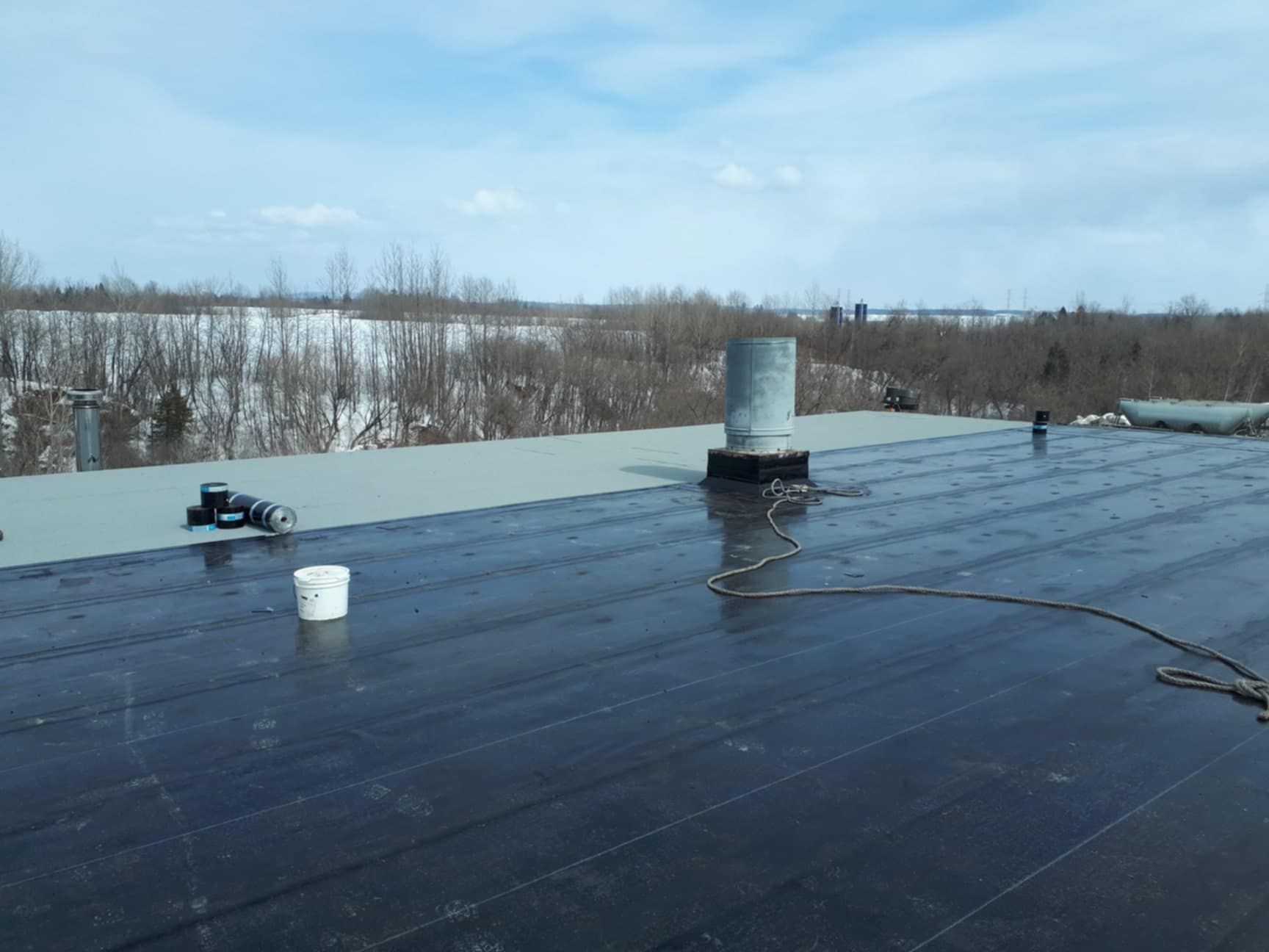 Toit plat noir avec un évent et un seau, paysage avec des arbres enneigés en arrière-plan.