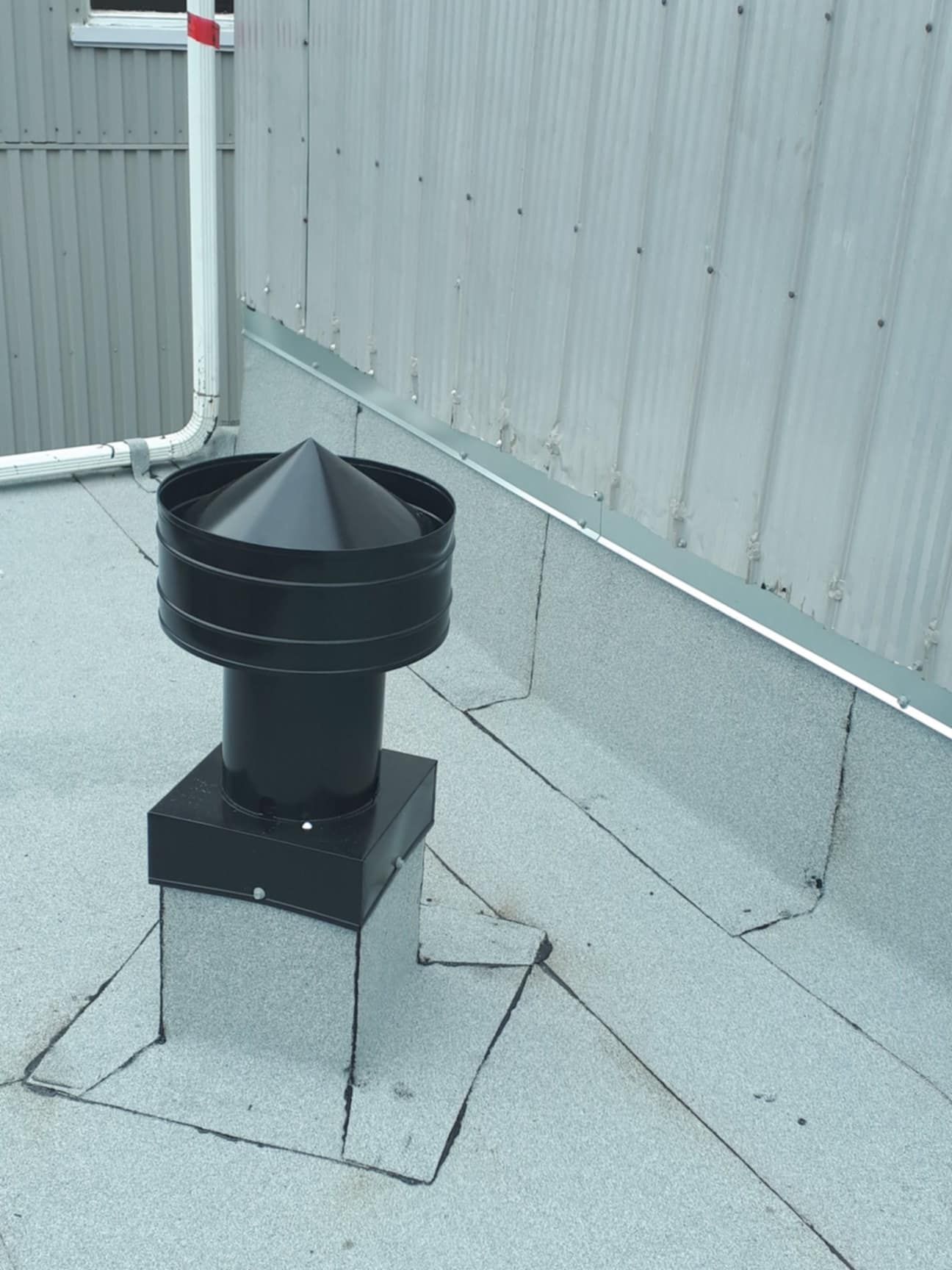 Chapeau de cheminée noir sur un toit plat et gris près d'un mur en métal ondulé et d'une gouttière blanche.