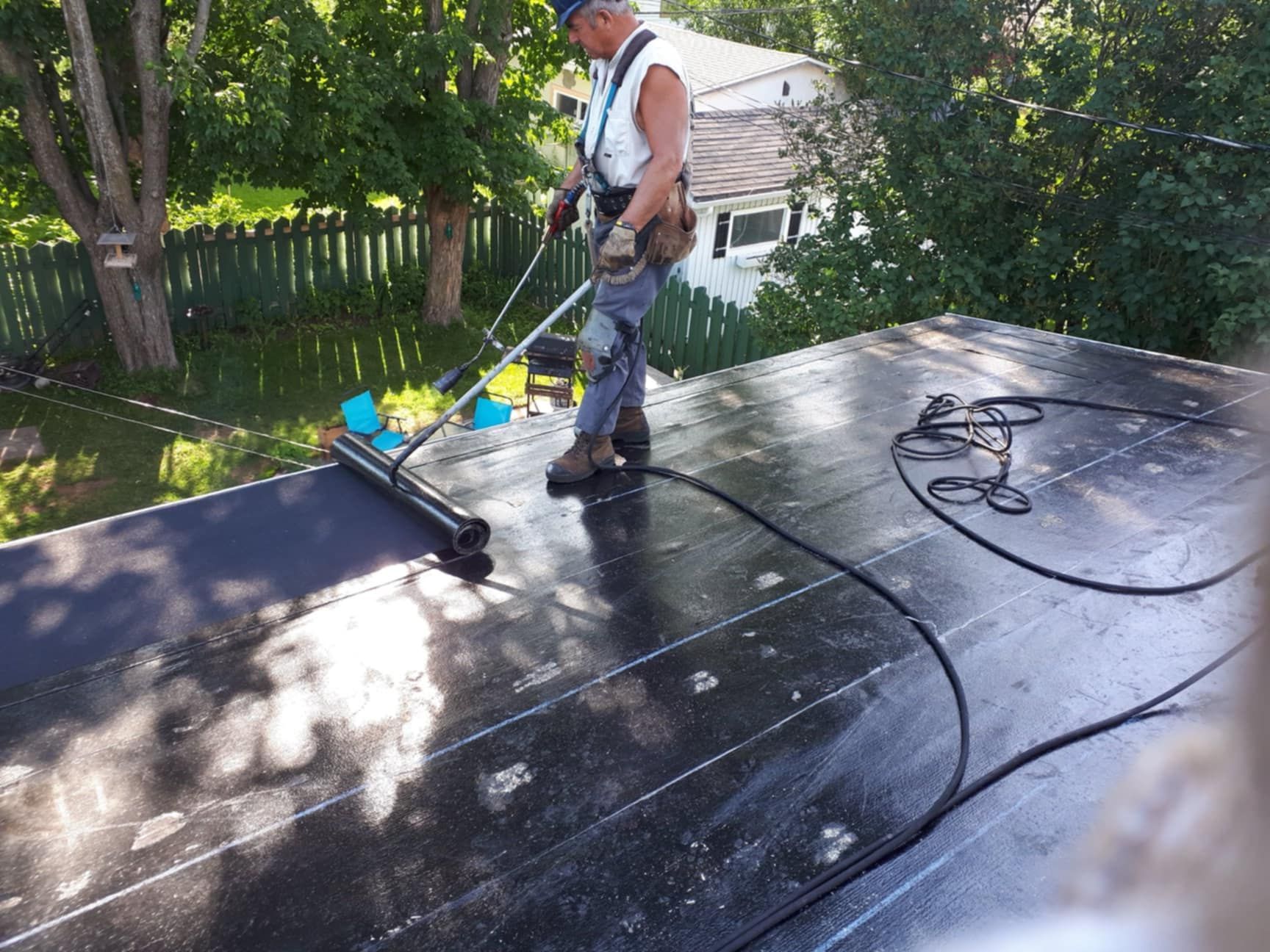 Couvreur sur un toit plat, appliquant du mastic avec un rouleau; portant un harnais de sécurité.