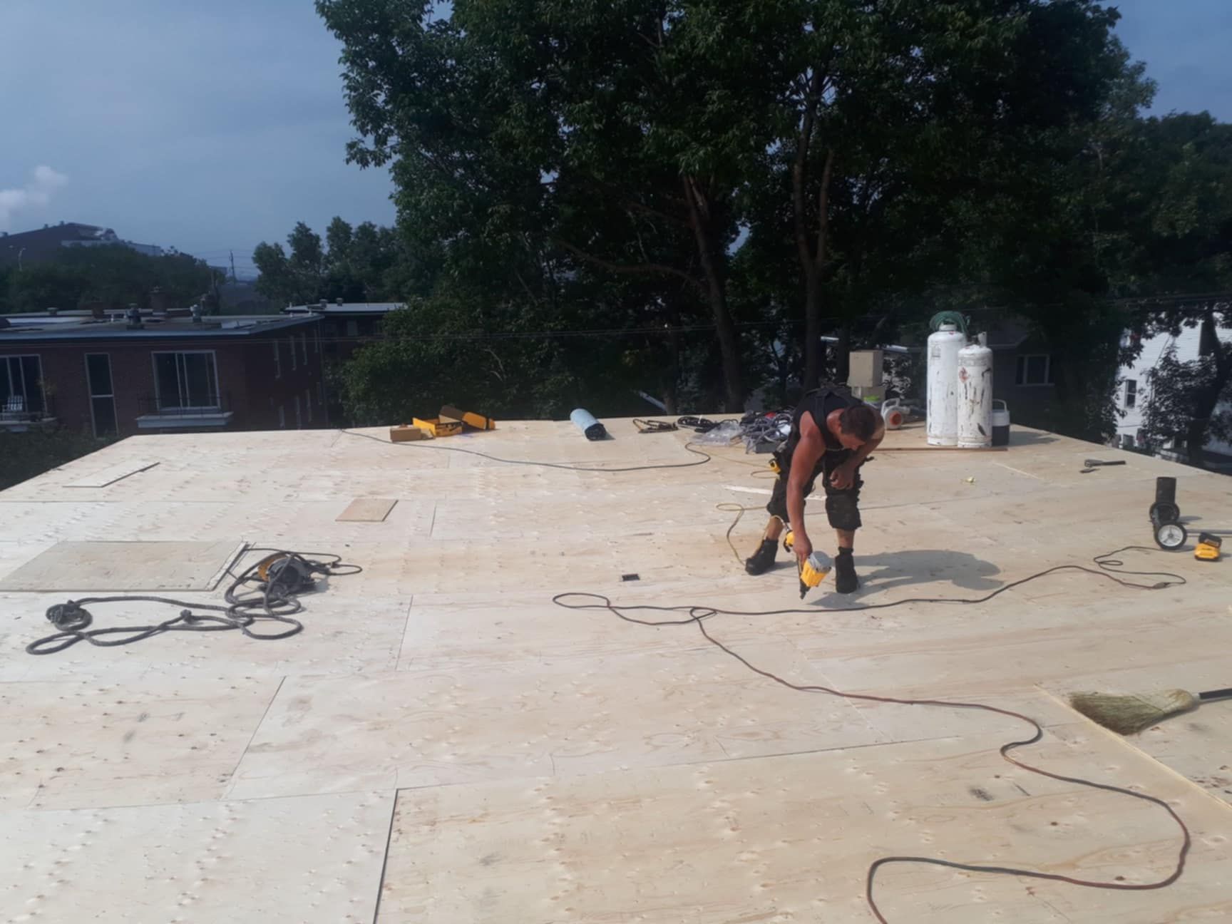 Un homme sur un toit utilisant un outil électrique, entouré d'outils et de matériaux ; arbres et bâtiments en arrière-plan.