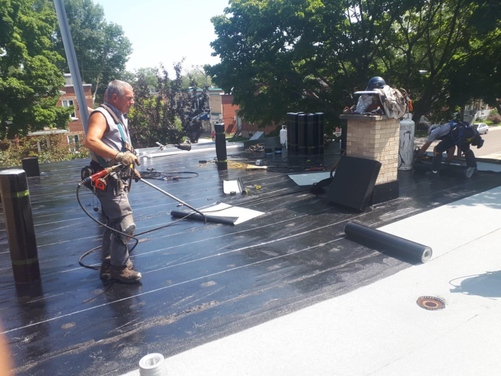 Couvreur utilisant un outil sur un toit noir, des arbres et une cheminée en arrière-plan par une journée ensoleillée.