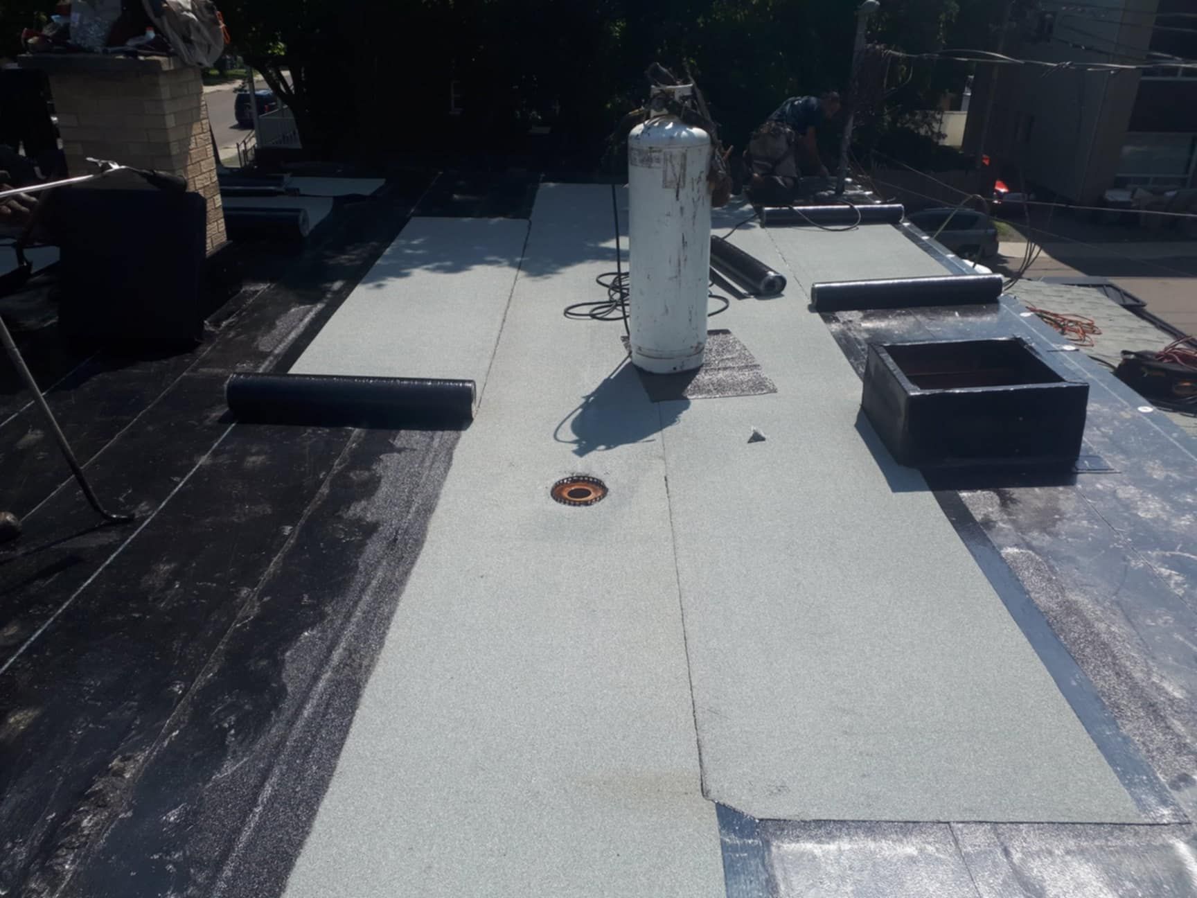 Toiture plate en travaux, avec matériaux de couverture blancs, gris et noirs. Réservoir de propane et tuyau d'aération présents.