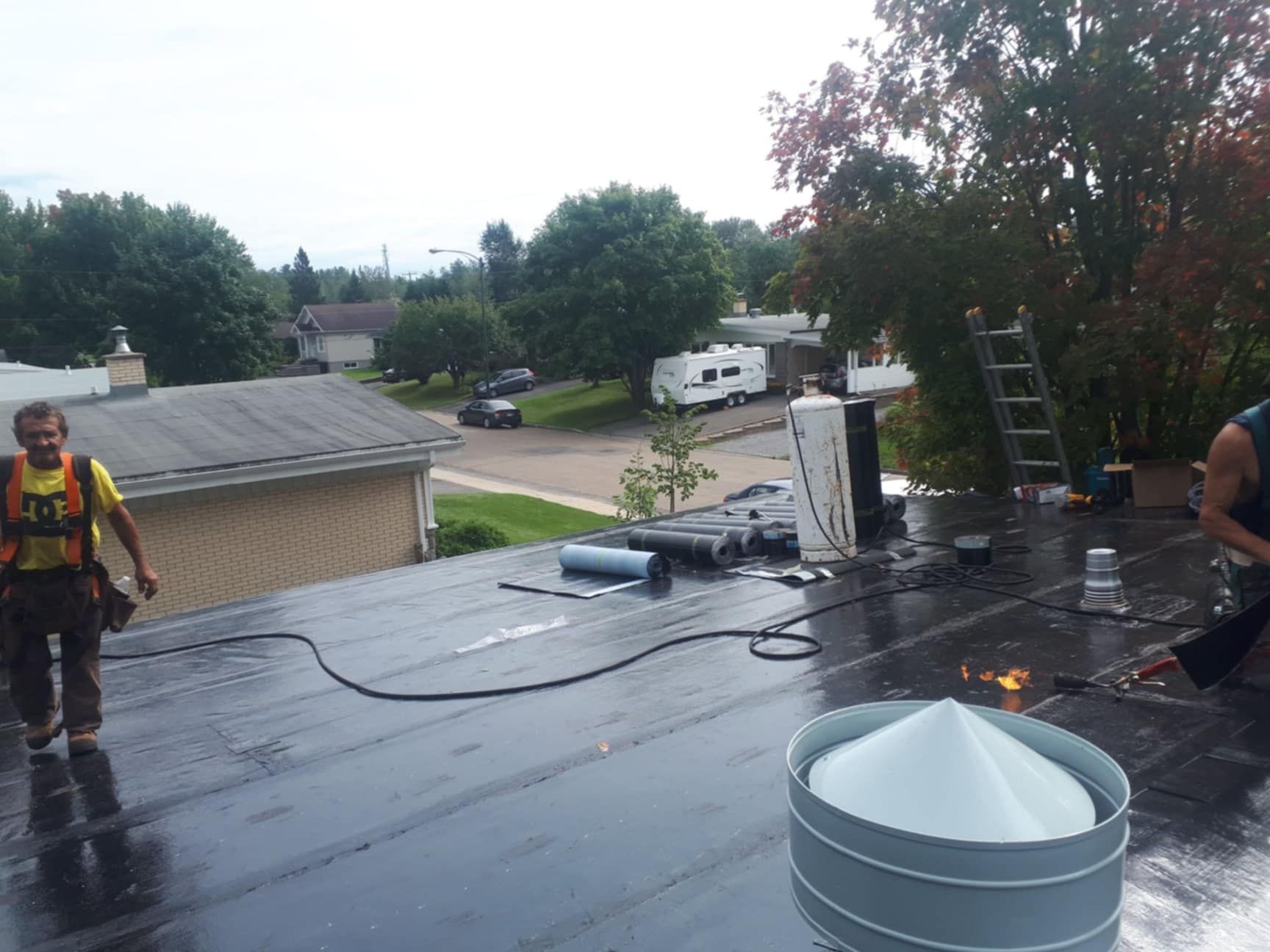 Couvreurs travaillant sur un toit plat, l'un d'eux utilisant un chalumeau. Arbres, maisons et un camping-car en arrière-plan.