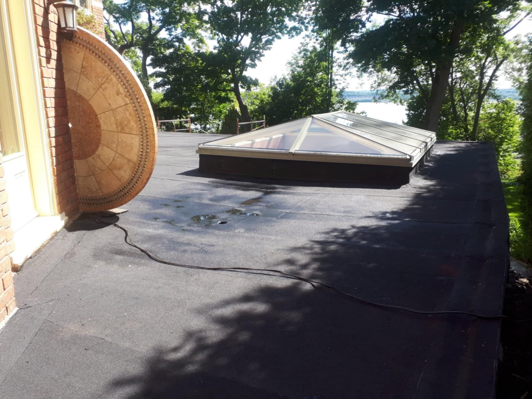 Un toit plat noir avec un puits de lumière et une table de patio extérieure ; des arbres et un plan d'eau en arrière-plan.