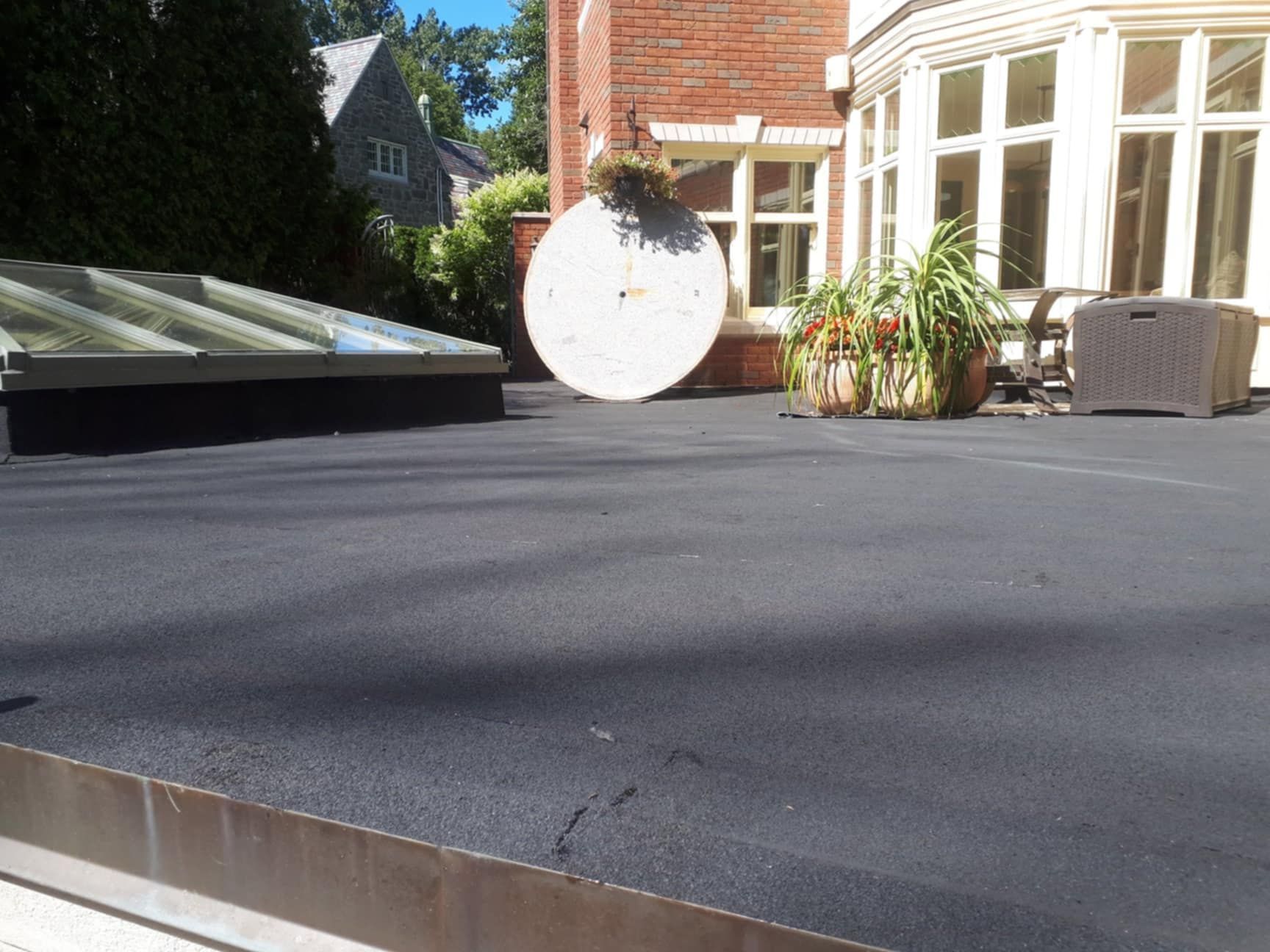 Toit plat noir avec puits de lumière, objet rond, plantes en pot et fenêtres dans un cadre extérieur ensoleillé.