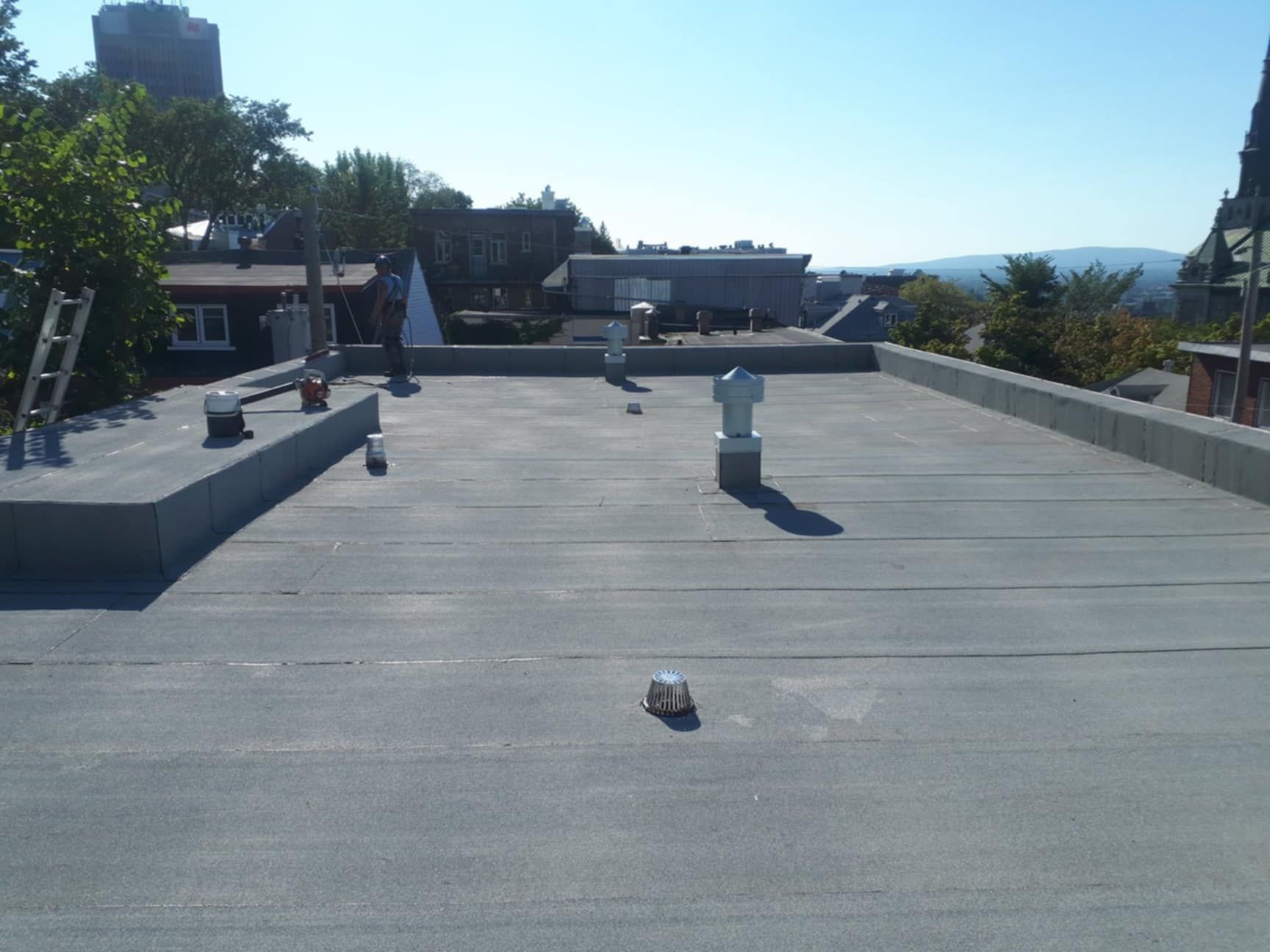 Toit plat avec plusieurs évents, bâtiments, arbres et une échelle par une journée ensoleillée.
